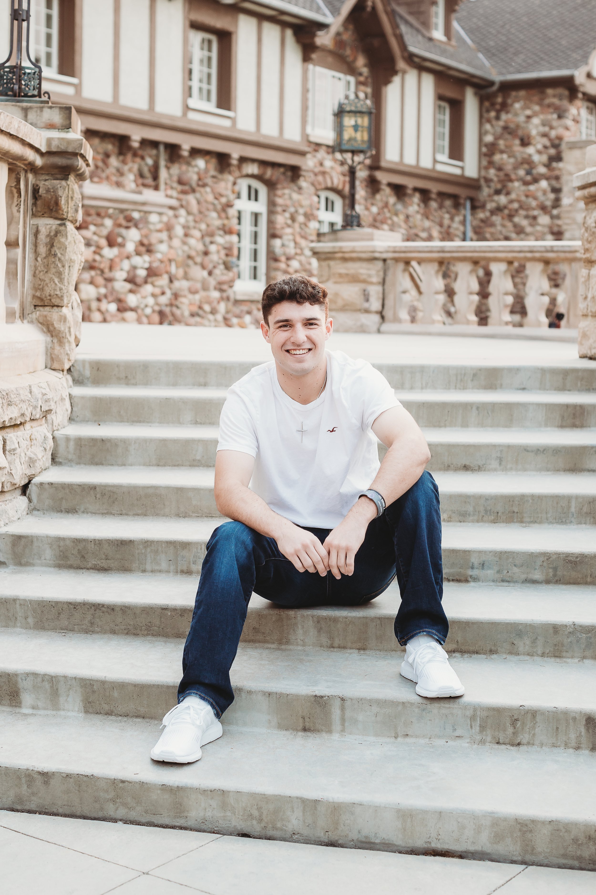A young man sitting on outdoor steps smiling at the camera in front of a stone and wood house with wrought iron lamps.
