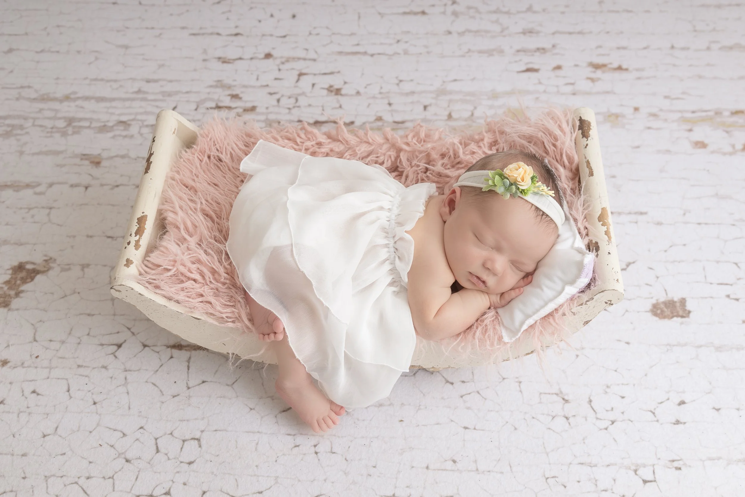 A sleeping baby girl lying on a small vintage white wooden bed with a pink fluffy blanket, wearing a white dress and a floral headband, on a distressed white wooden floor.