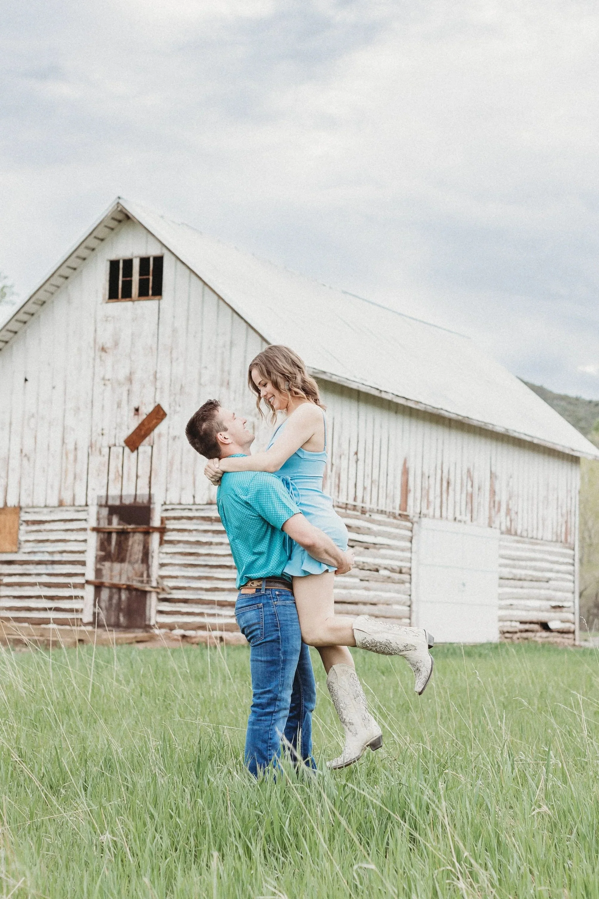 A couple standing in a grassy field, with the man lifting the woman, both smiling and looking at each other, in front of an old wooden barn.
