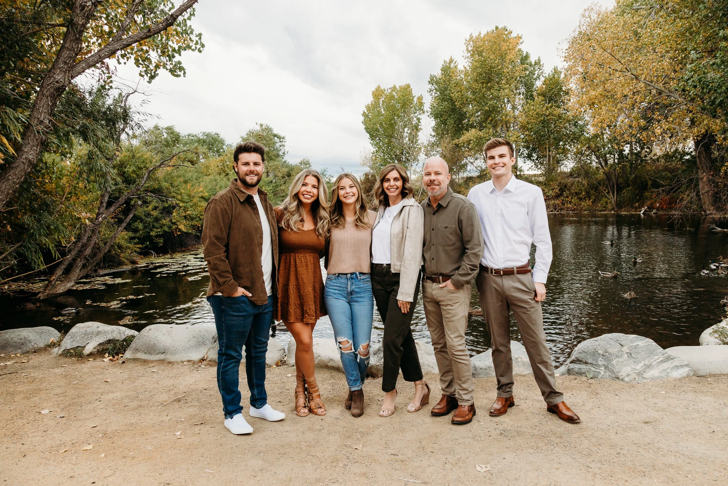 Group of six people standing on dirt path by water with trees and fall foliage in background.