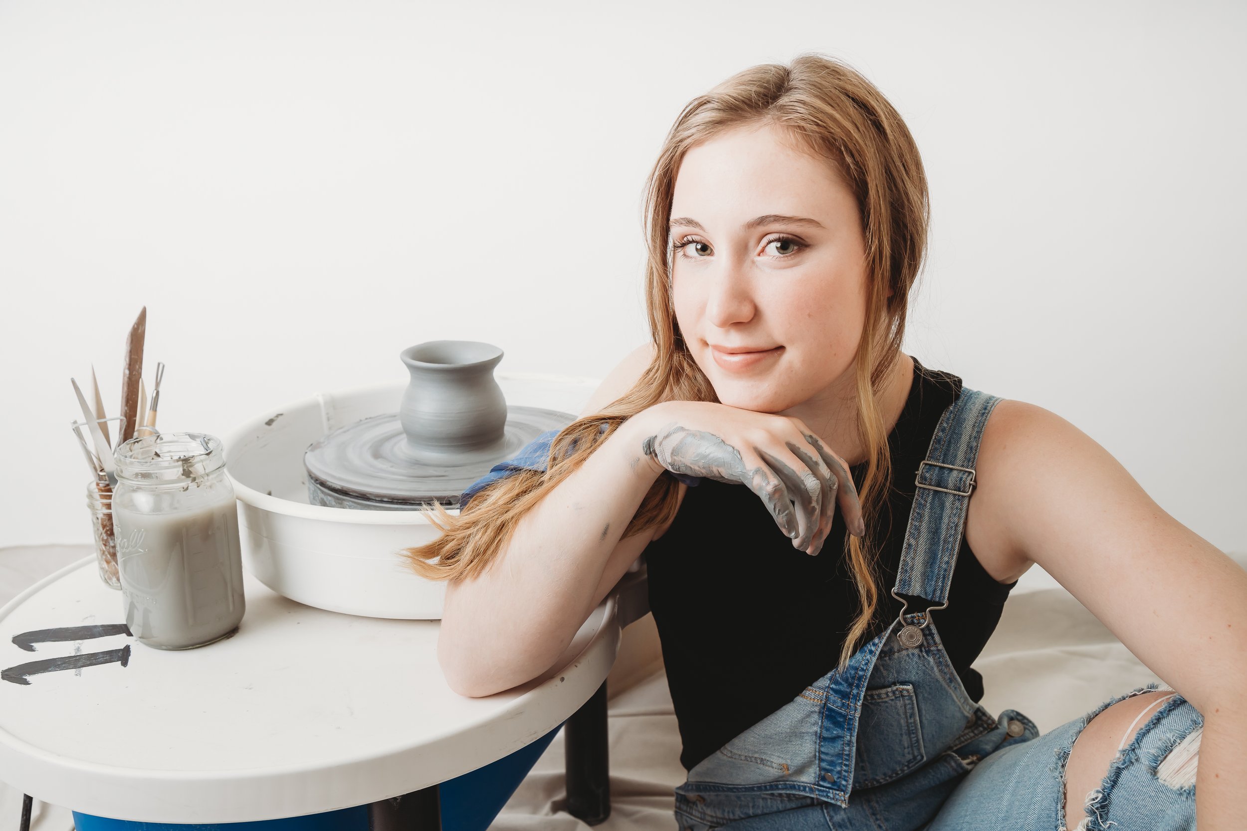 Young woman with mud-streaked hands sitting at a pottery wheel with a partially shaped clay vase, smiling and resting her chin on her hand, in a bright studio.