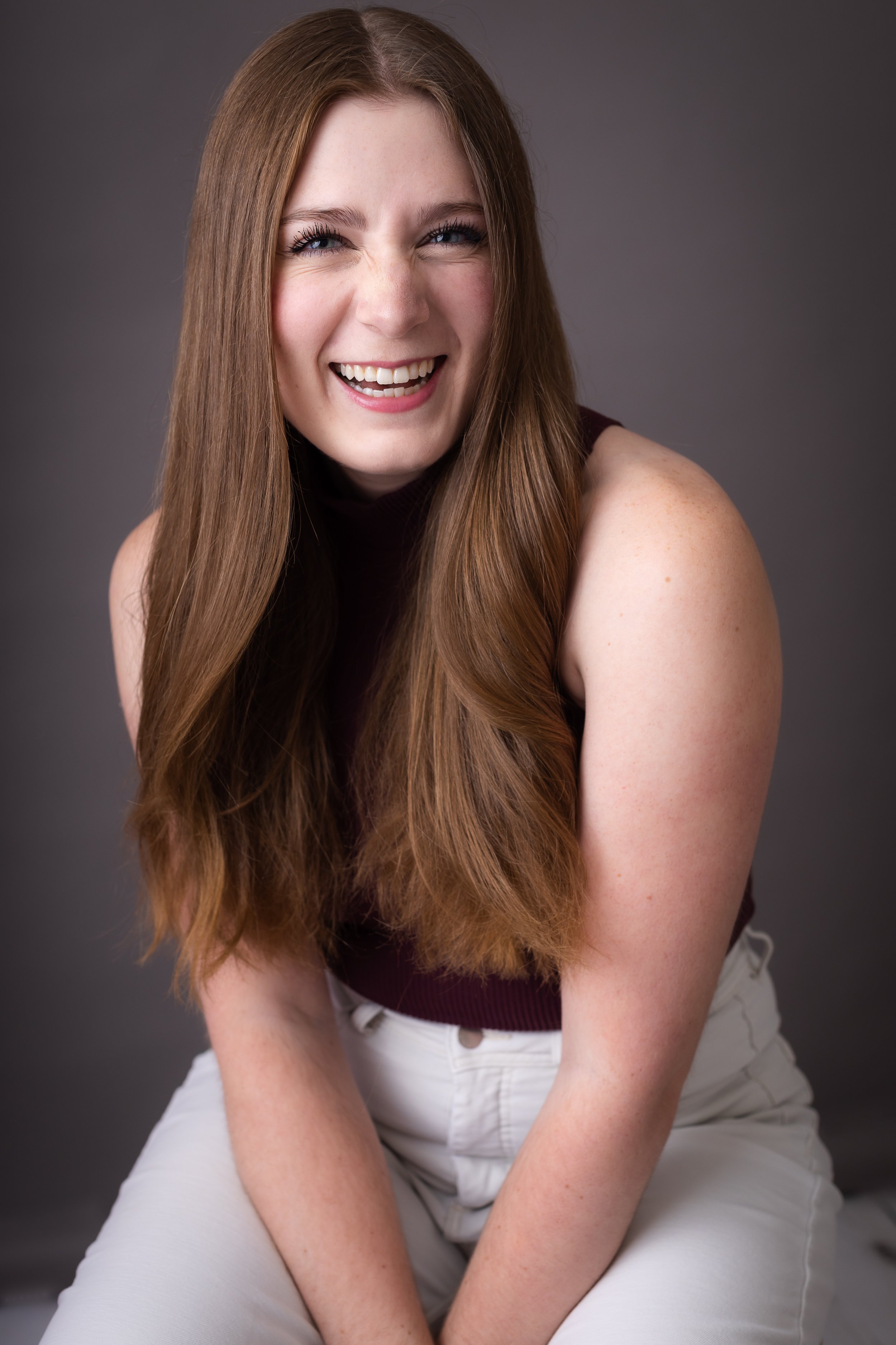 A young woman with long, wavy red hair, smiling and wearing a sleeveless dark maroon top and white pants against a neutral gray background.