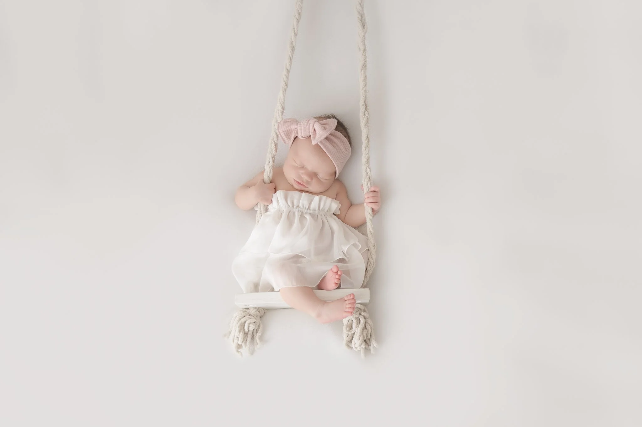 A sleeping baby girl on a white swing with knotted ropes, wearing a pink headband with a bow and a white dress, lying on a white background.