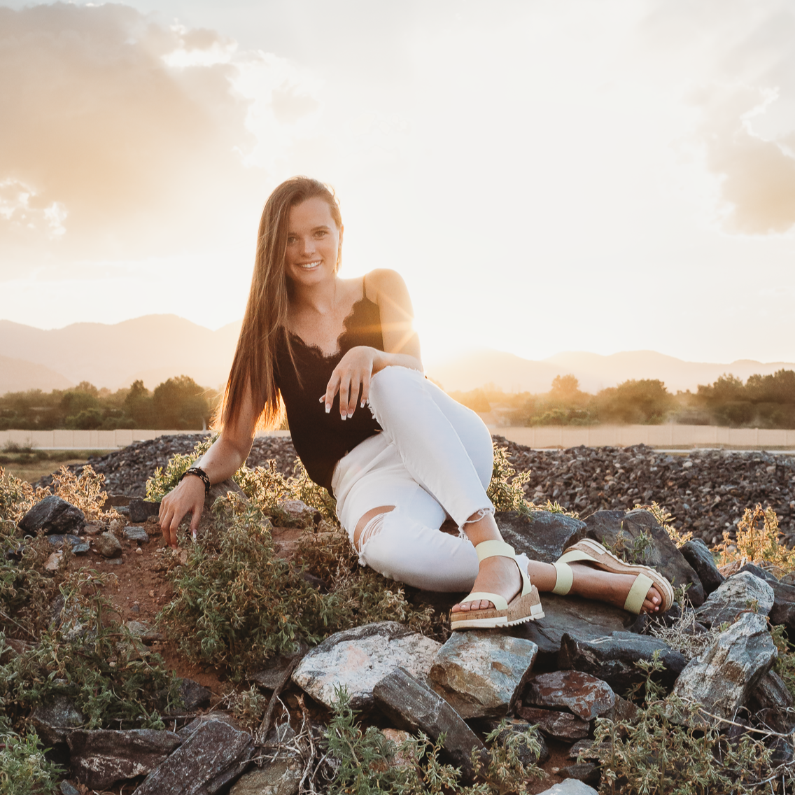 A woman sitting on rocks outdoors during sunset, smiling, with mountains and clouds in the background.