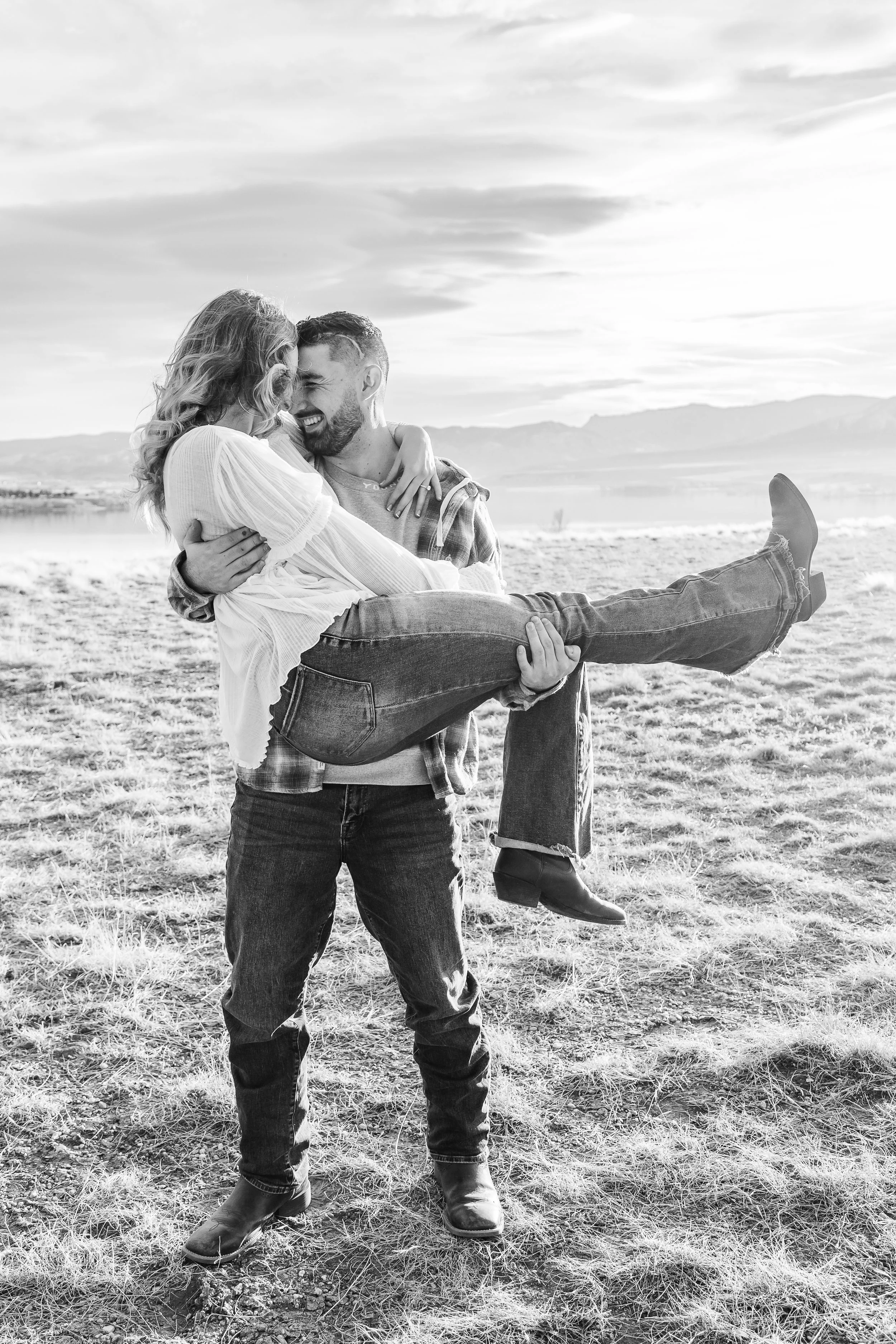 A man holding a woman in his arms on a field with a landscape and mountains in the background, both smiling and looking at each other.