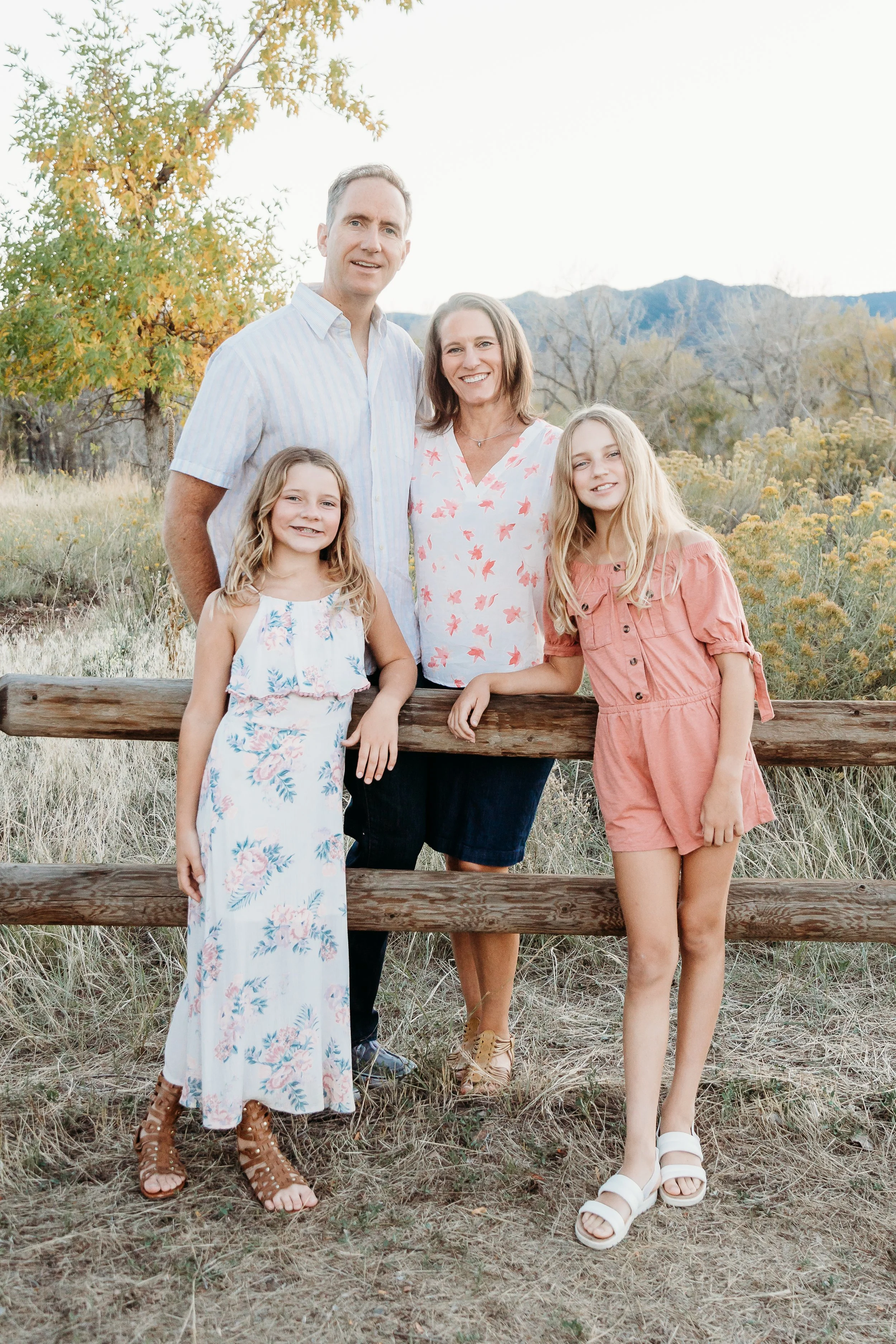 A family of four standing outdoors near a wooden fence, with mountains and trees in the background, during late afternoon or early evening.