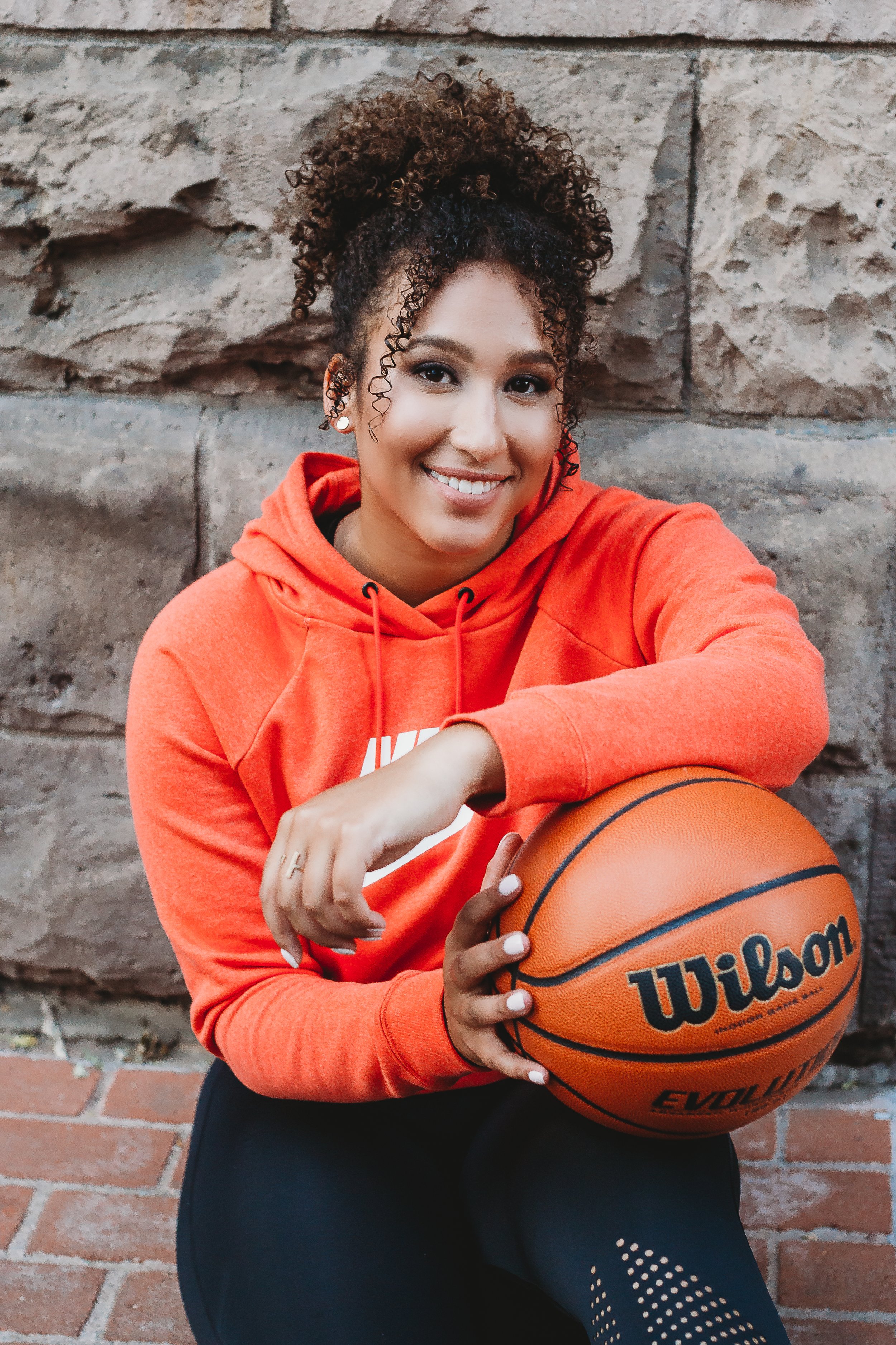 A young woman with curly hair, wearing an orange Nike hoodie, smiling and holding a Wilson basketball, sitting against a stone wall.