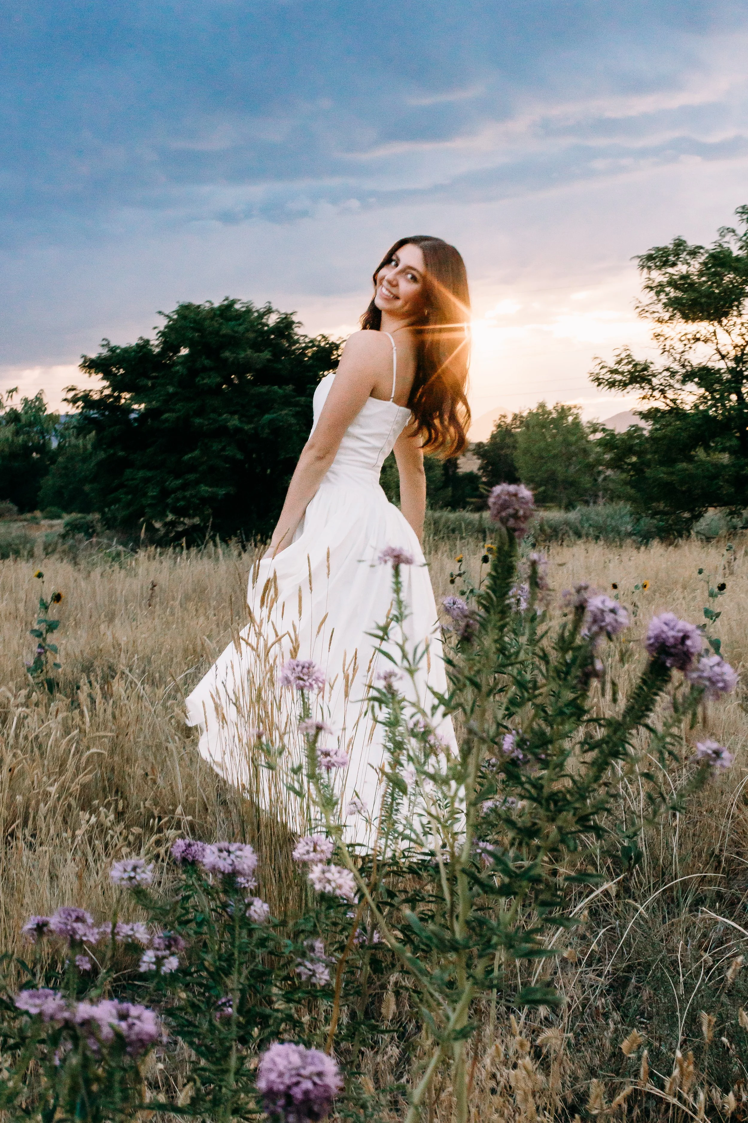 Woman in a white dress standing in a field of tall grass and purple flowers during sunset, smiling.