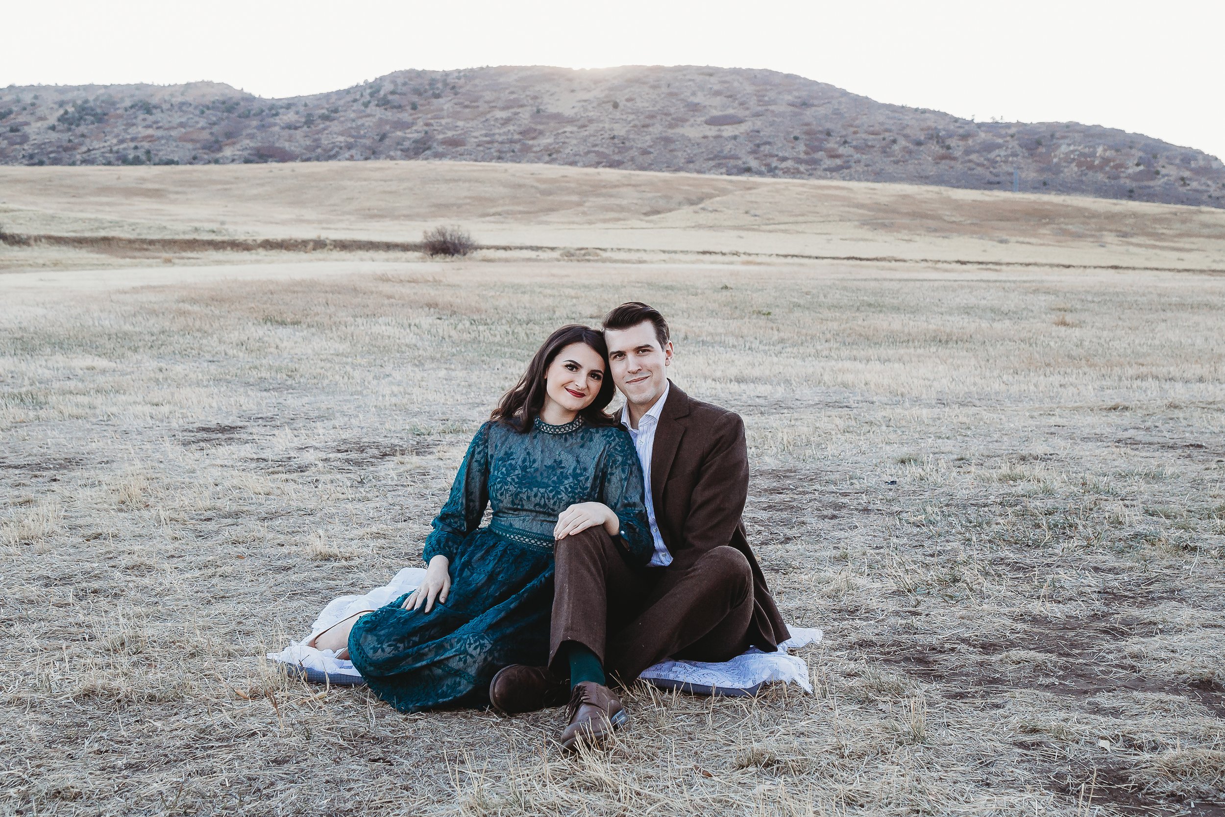 A couple sits on a blanket in an open field with hills in the background, smiling at the camera.