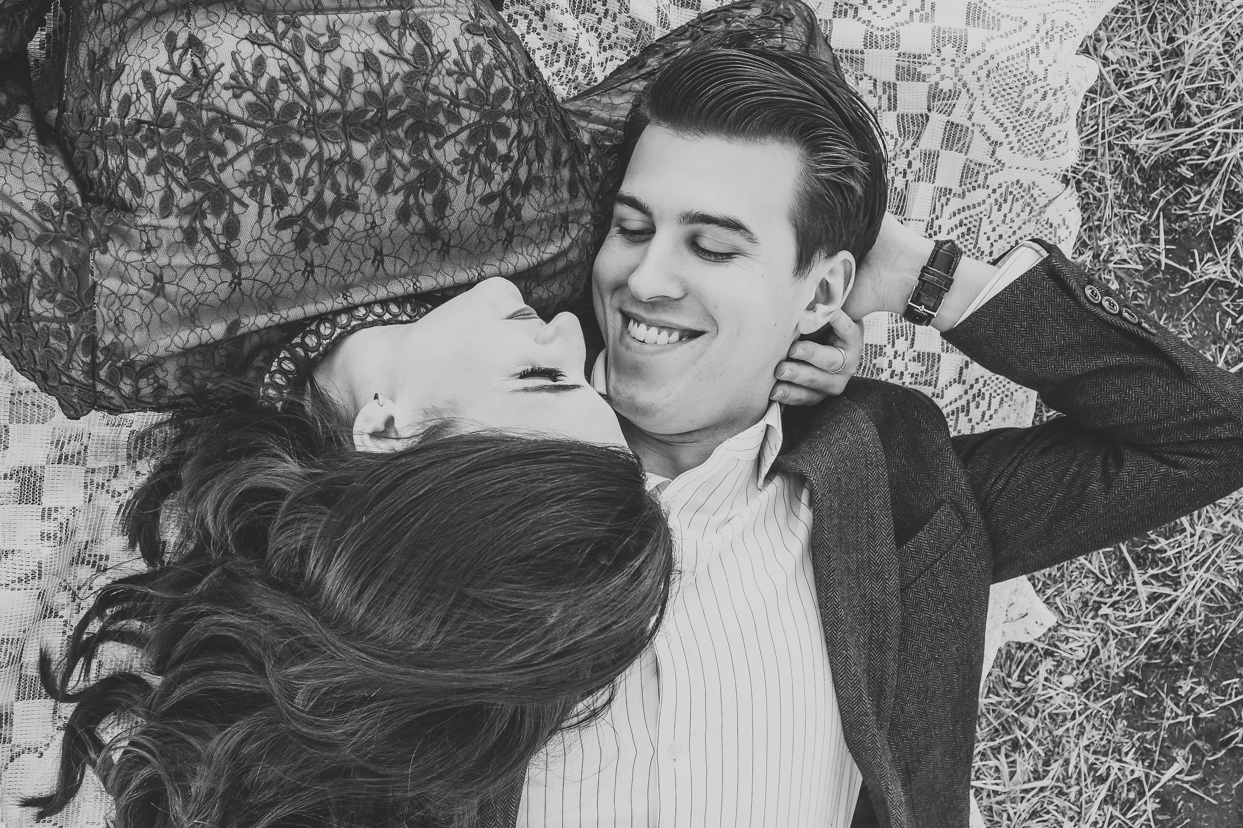 A young man and woman lie on a checkered blanket outside on the grass, smiling at each other with their heads close together. The woman has long hair, wears earrings, and a lace top. The man has short, neatly styled hair, and is dressed in a suit jac