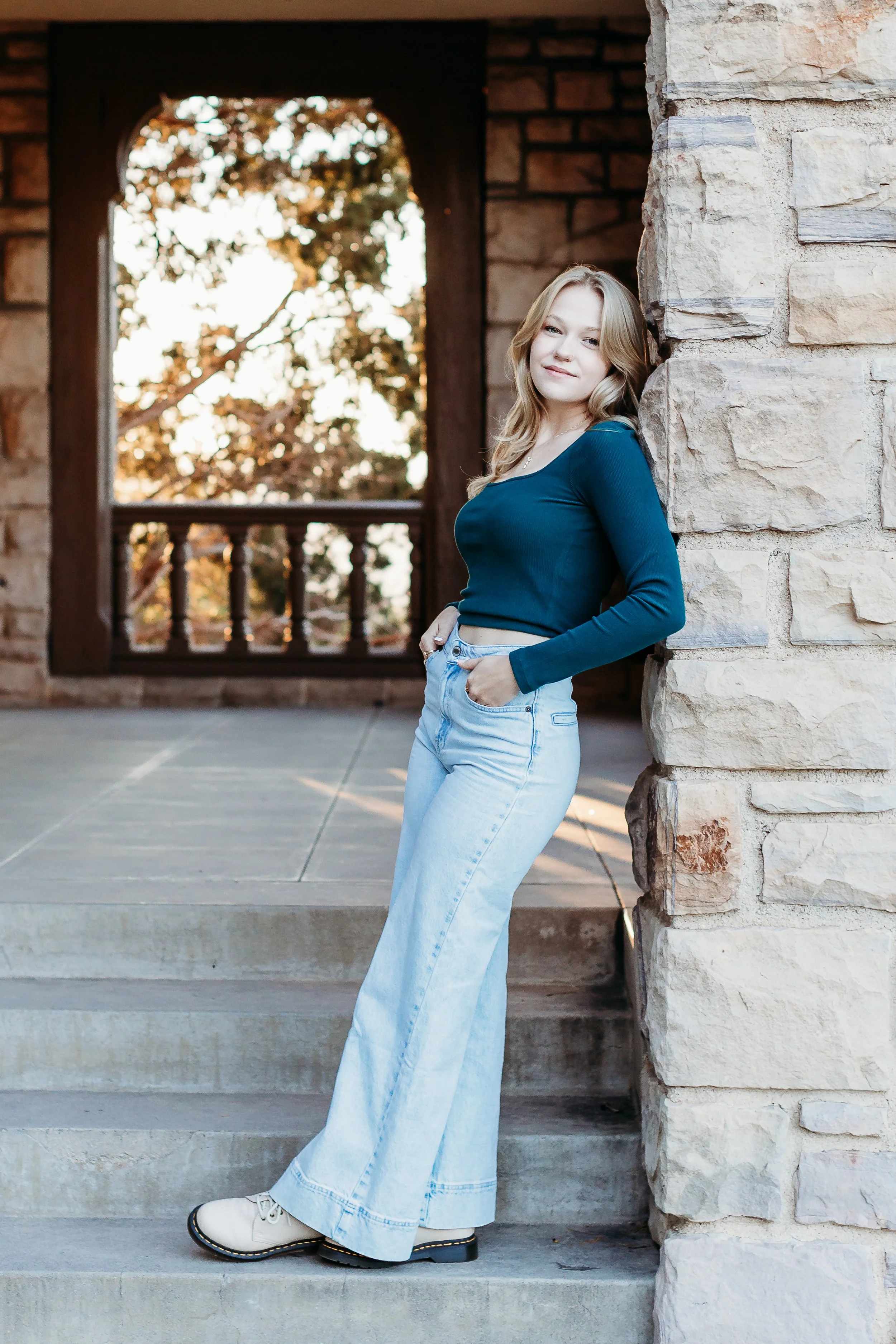 A young woman with light brown hair in loose waves, wearing a teal long-sleeve crop top, light blue wide-leg jeans, and beige shoes, leaning against a stone wall on steps outside a brick building with a wooden balcony, during golden hour.