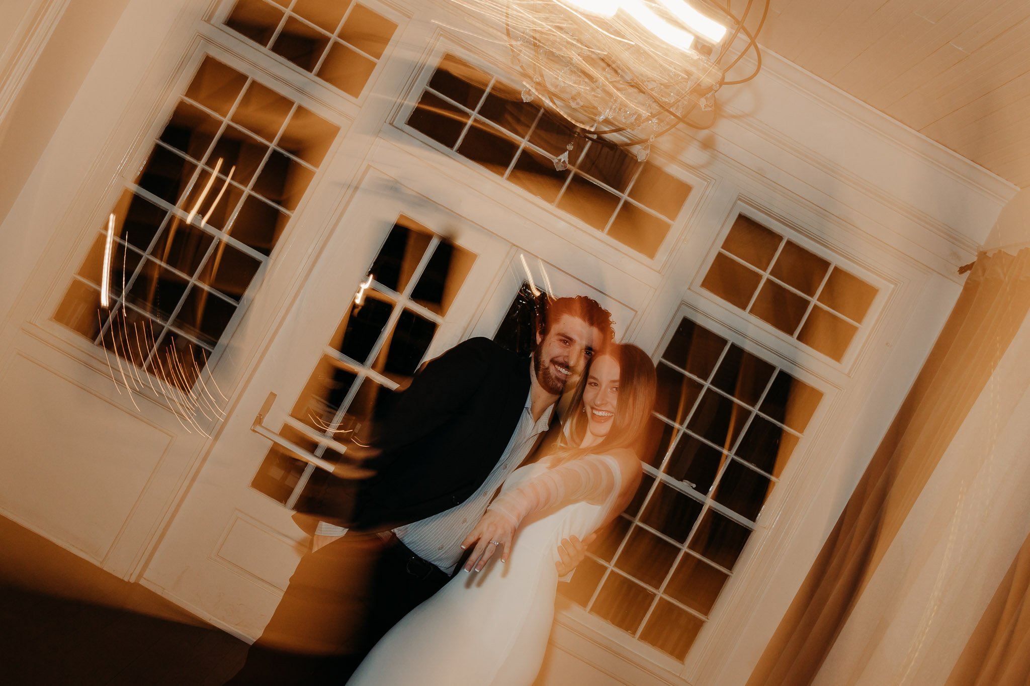 A happy couple in formal attire standing close together and smiling at a party or wedding reception in a room with large windows and curtains.