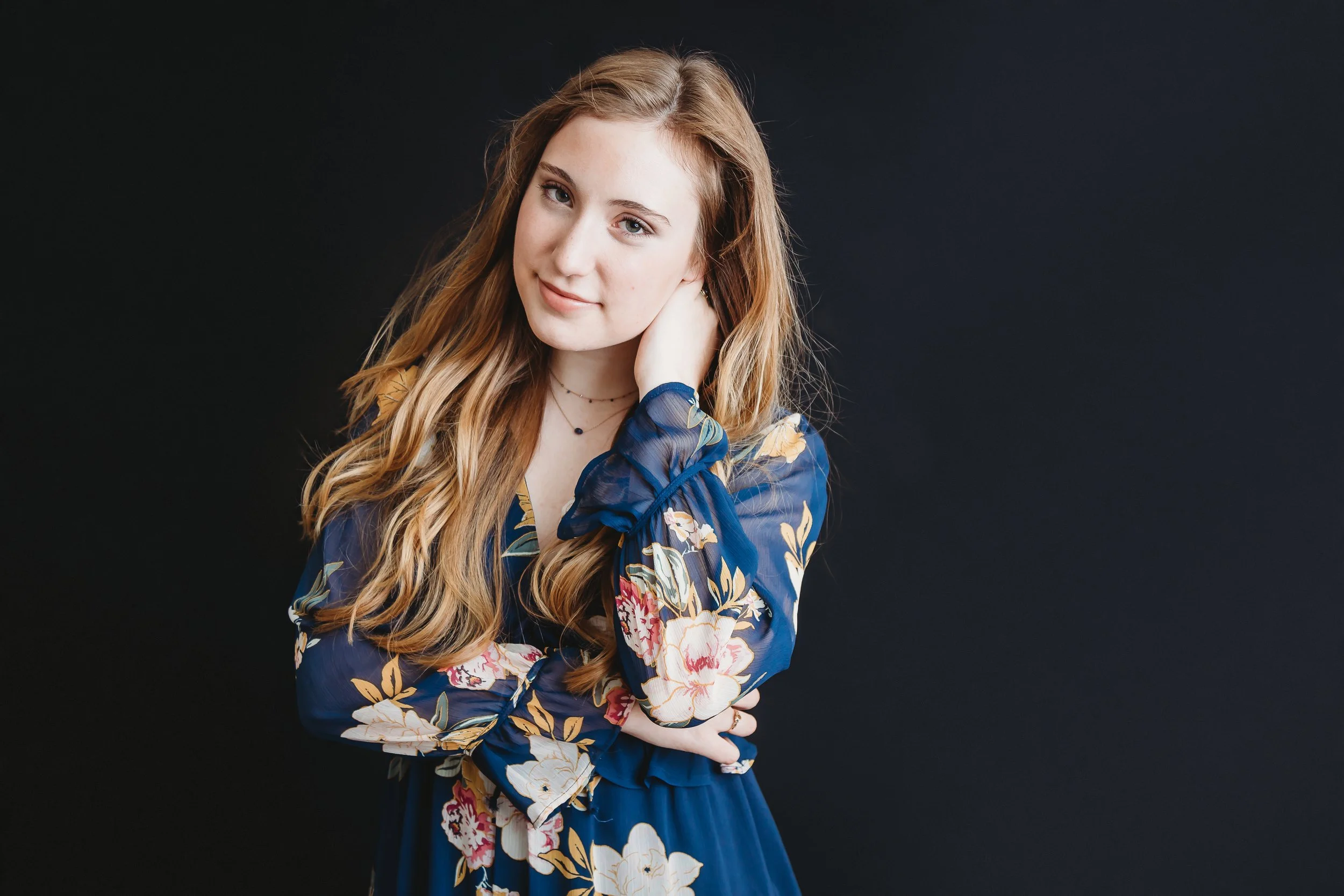 A young woman with long, wavy, light brown hair, wearing a navy blue floral dress, posing with one hand touching her neck against a dark background.