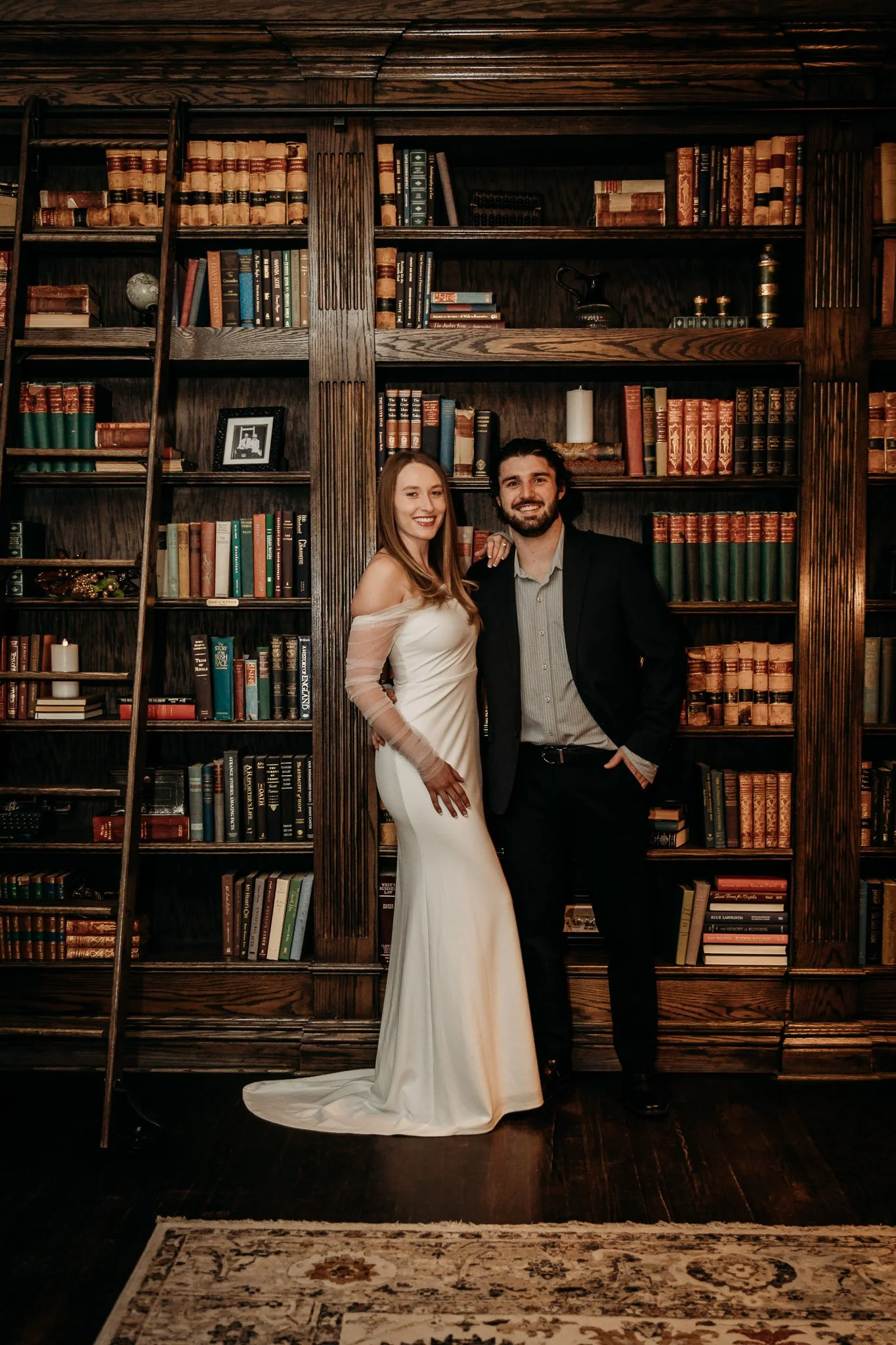 A couple dressed in formal attire posing in front of a large wooden bookshelf filled with books, candles, and decorative items, in a cozy, dimly lit room.