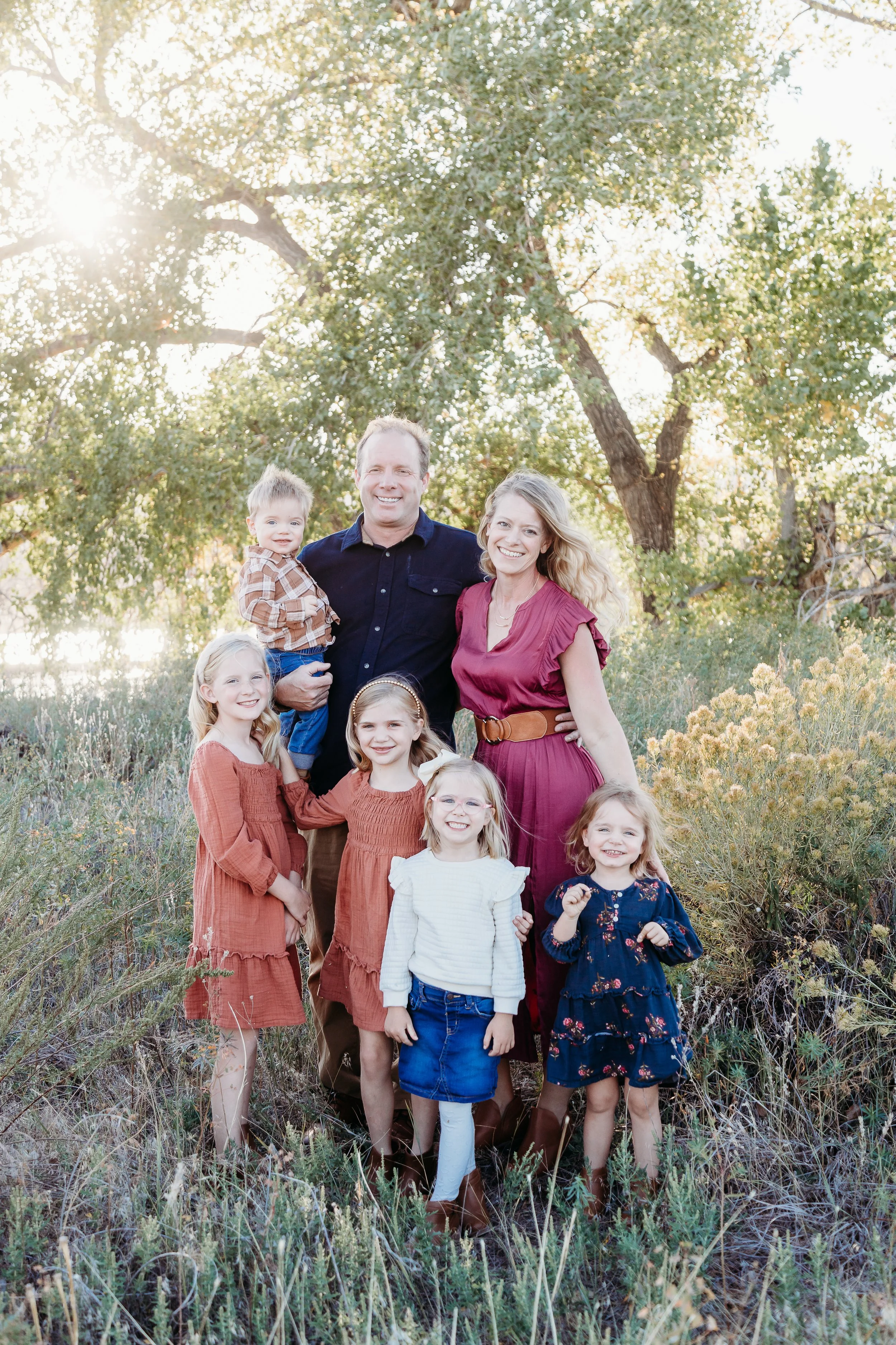 Family of seven standing outdoors in a natural setting with sunlight filtering through trees, smiling at the camera.