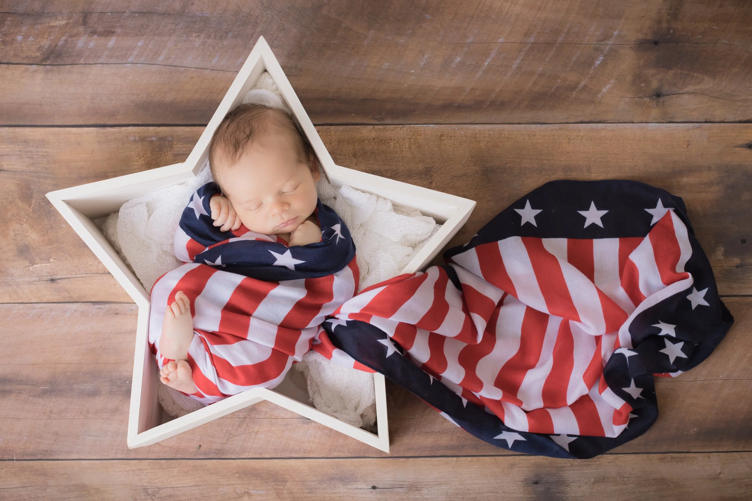 A newborn baby sleeping in a star-shaped wooden box, wrapped in a red, white, and blue American flag blanket, on a wooden floor.