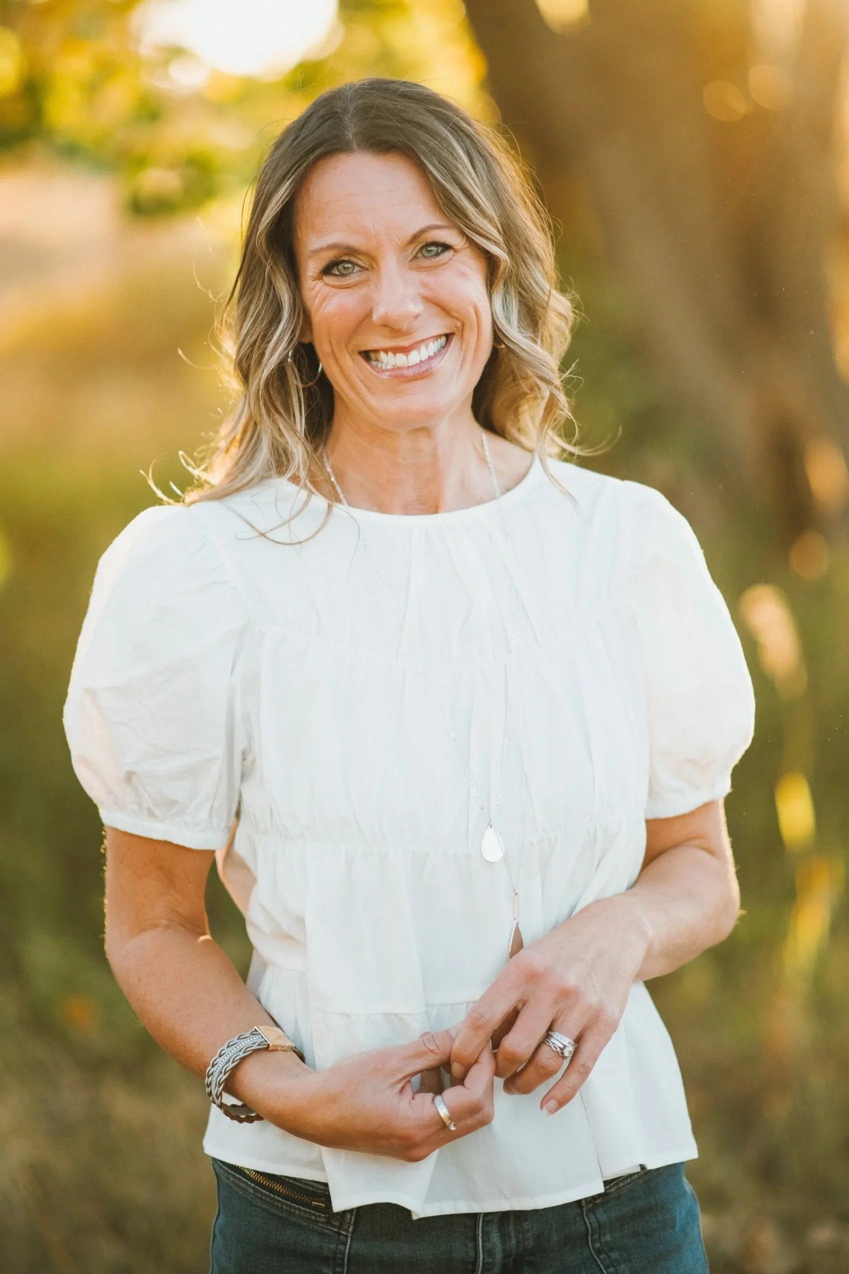 A woman with shoulder-length wavy hair, smiling, standing outdoors near a body of water with trees in the background, wearing a white blouse with puffed sleeves and jewelry.