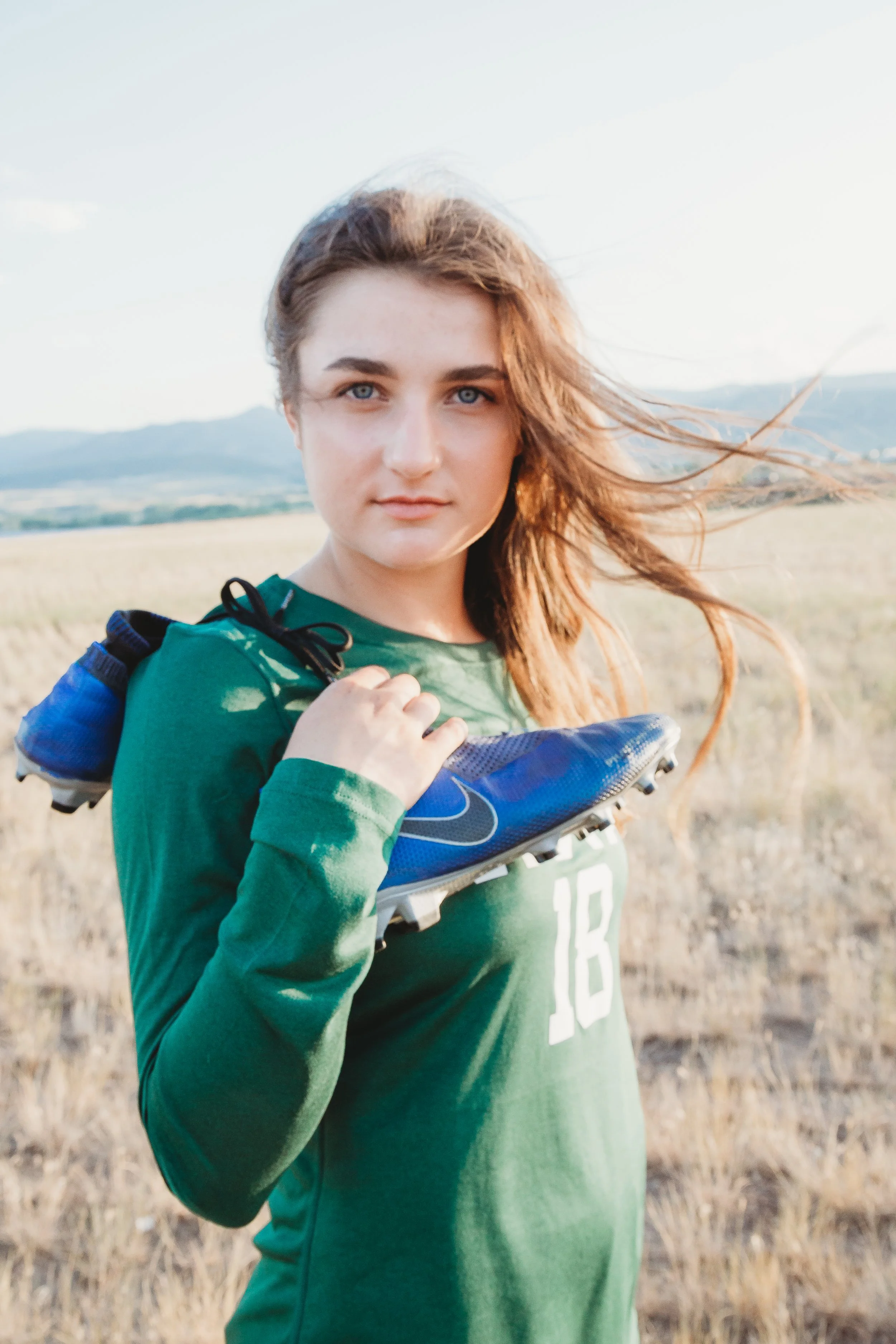 Young woman with long hair standing outdoors in a grassy field, holding a blue soccer shoe over her shoulder, wearing a green sports jersey with the number 18, with mountains in the background during daytime.
