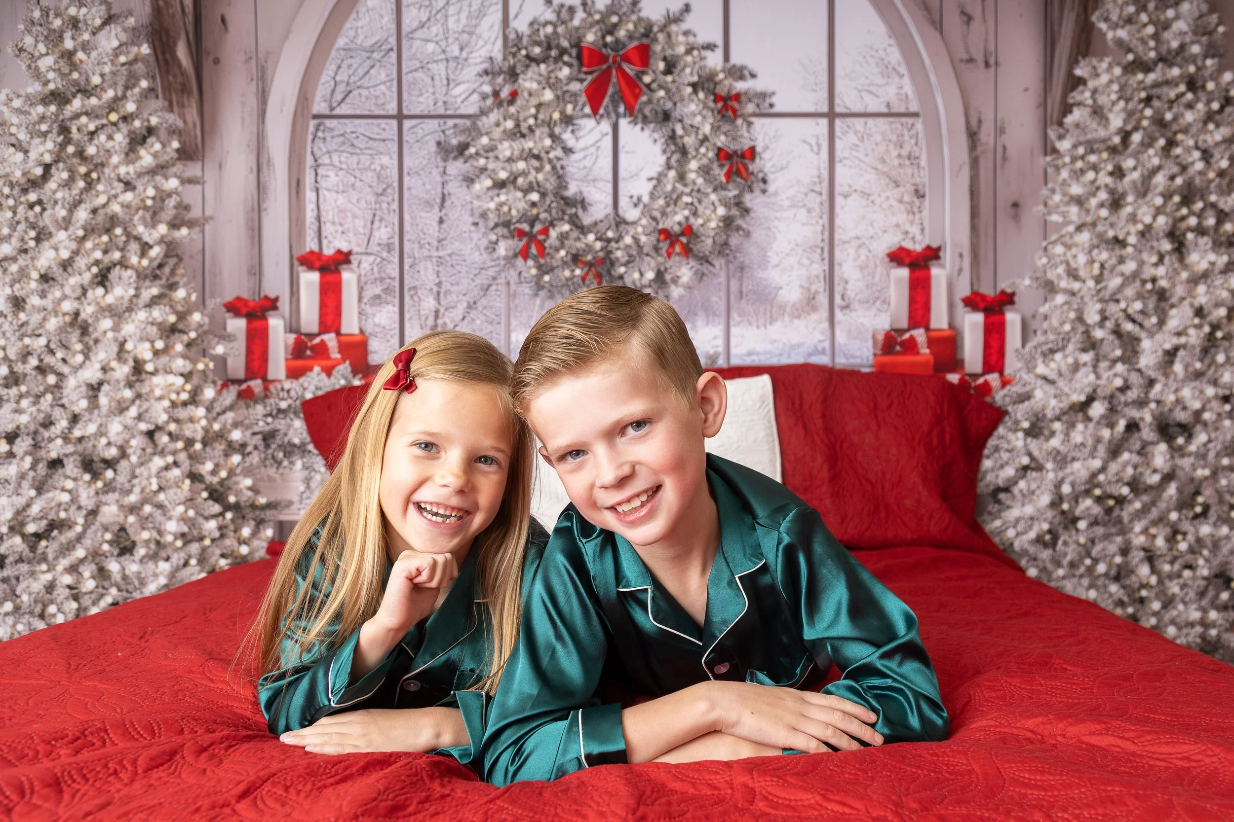 Two children, a girl and a boy, lying on a red bed in a Christmas setting with decorated trees, a large window, a wreath, and wrapped presents.