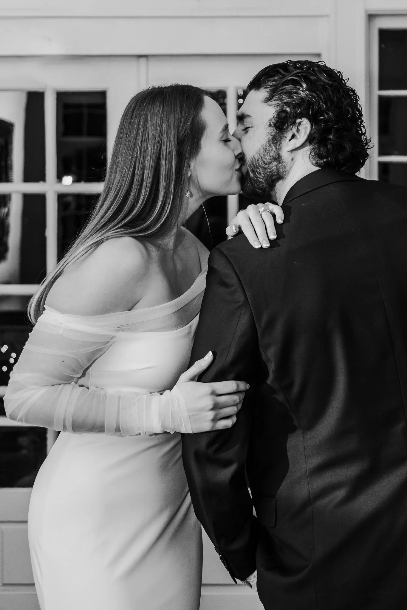 A black-and-white photo of a woman in an off-shoulder gown and a man in a suit sharing a kiss at an indoor event.