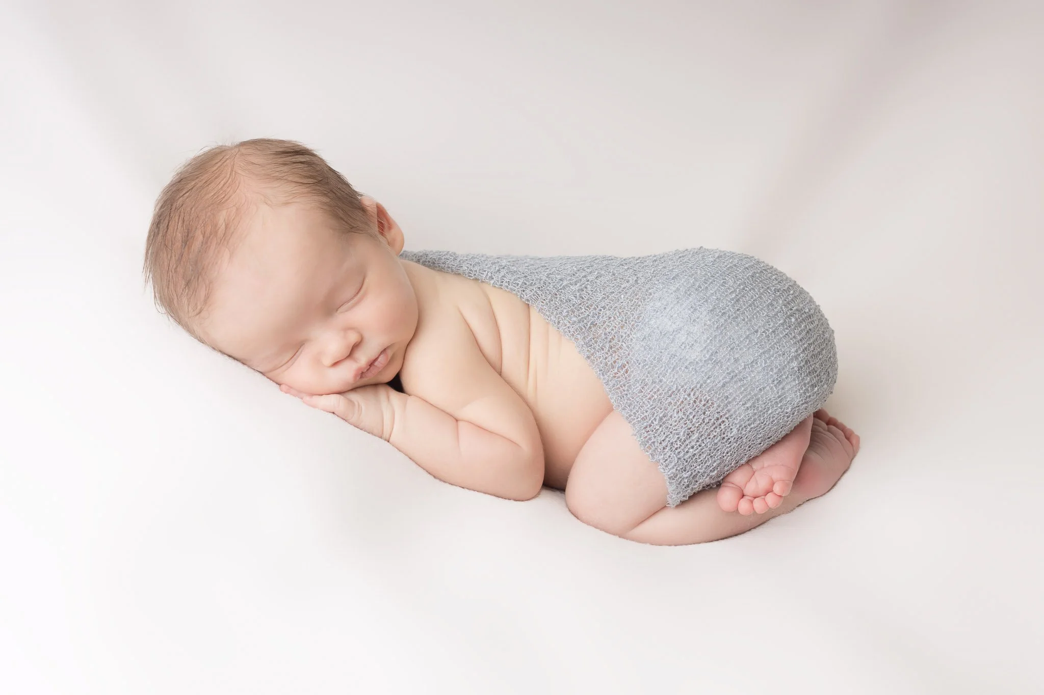 Softly sleeping newborn baby curled up on a white surface, wrapped in a gray knitted blanket, with one hand under the cheek and the other resting near the face.