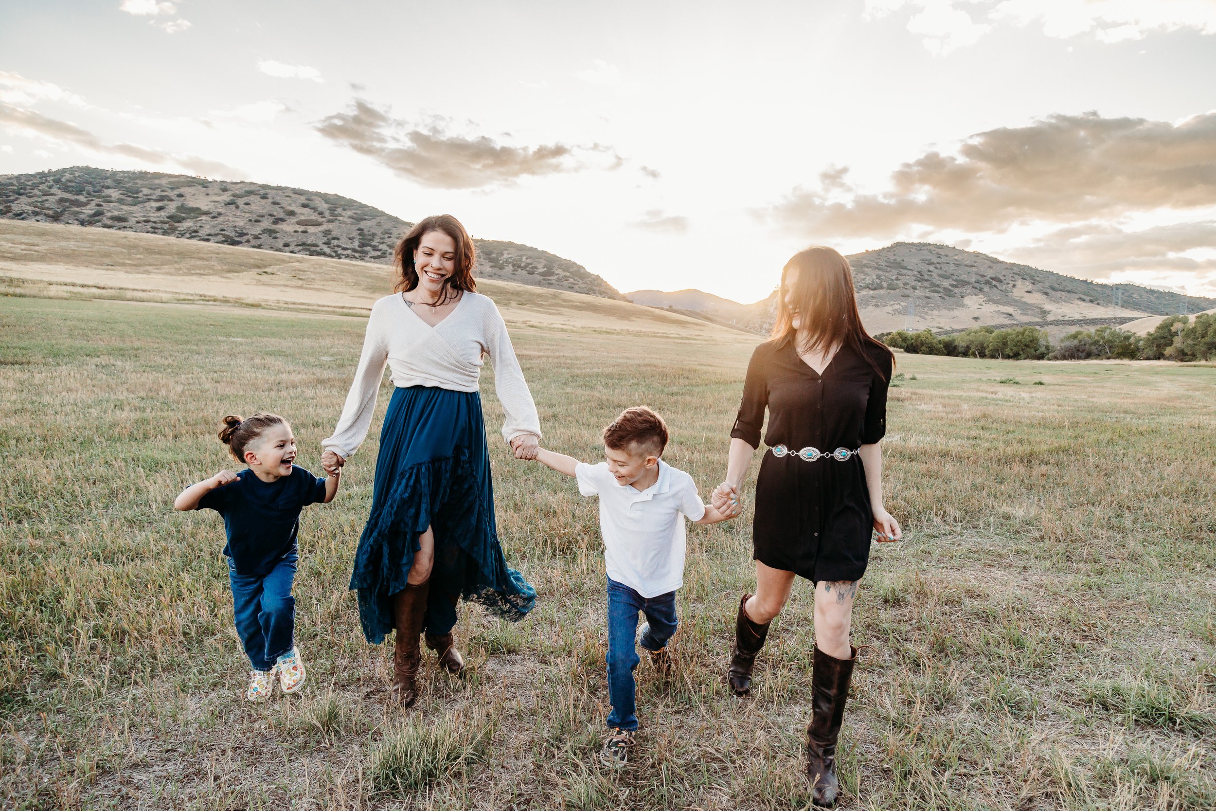A mother and her three children holding hands and running across a grassy field during sunset, with hills and a partly cloudy sky in the background.