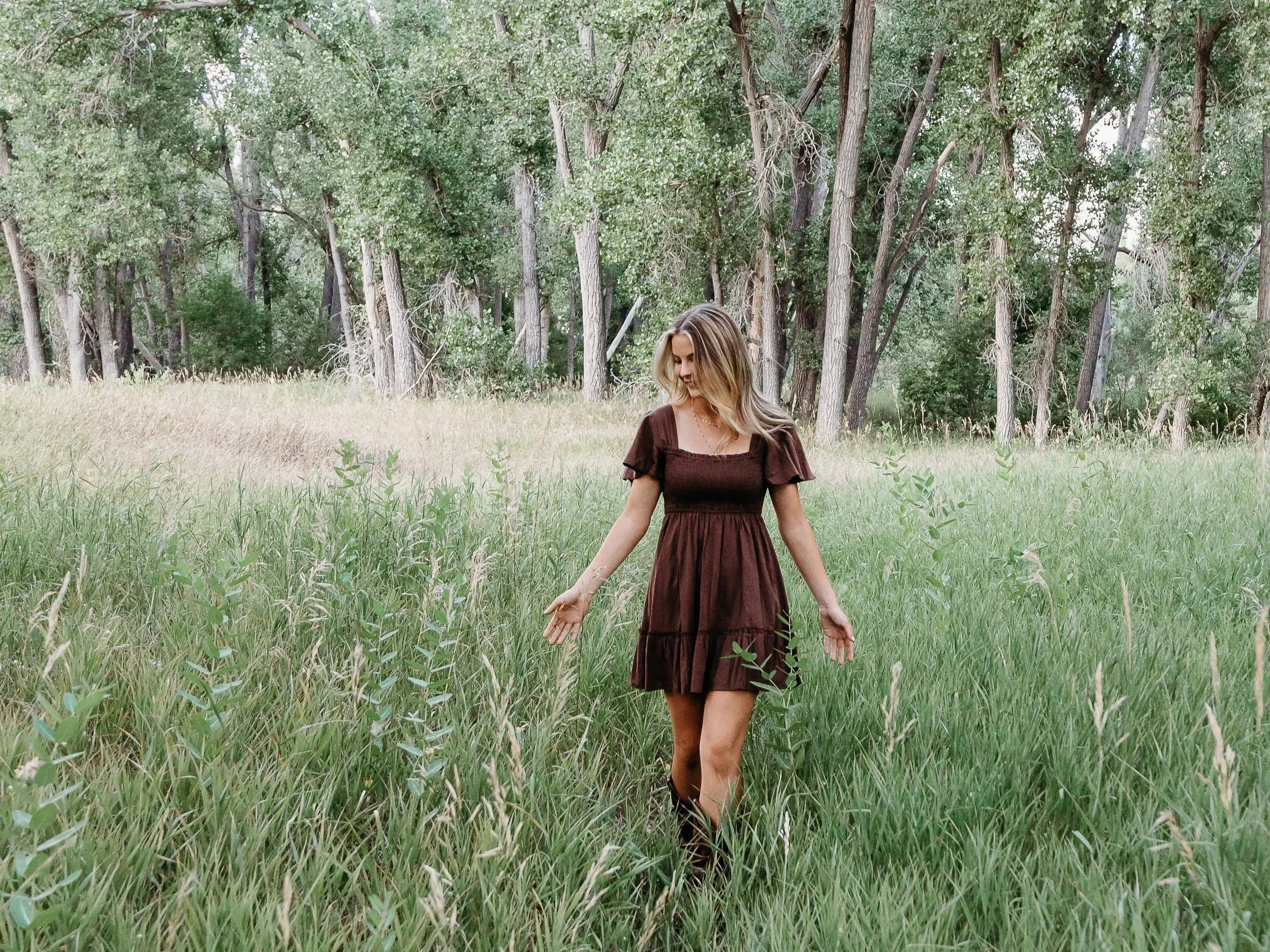 A woman in a dark brown dress walking through tall green grass in a field with trees in the background.