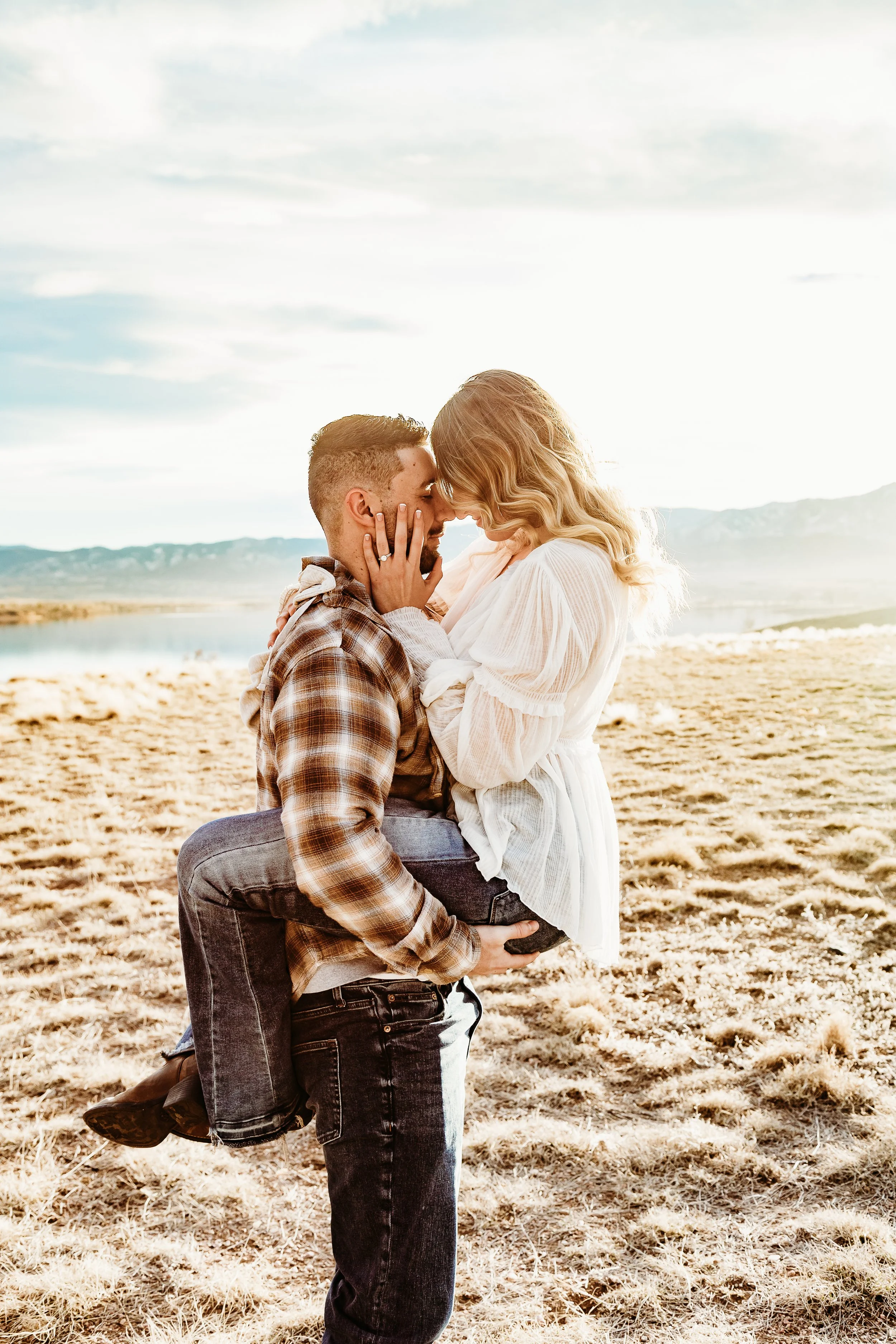 A man holding a woman in his arms on a grassy, outdoor area near a body of water, with mountains and a bright sky in the background, as they lean in close with their foreheads touching.