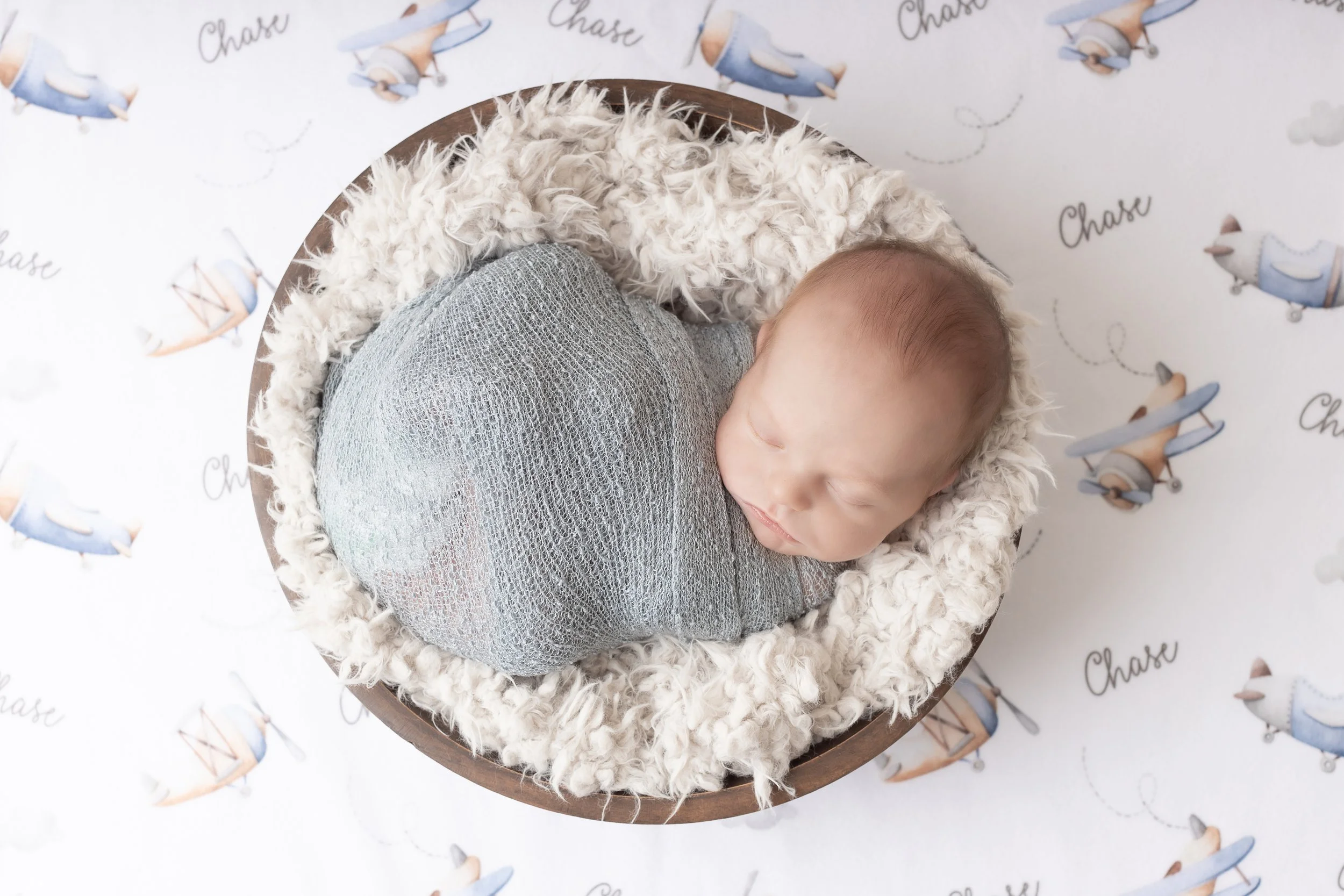 A newborn baby sleeping in a round basket lined with a fluffy white blanket, wrapped in a gray mesh cloth, on a patterned floor with airplanes and the word 'Chase'.