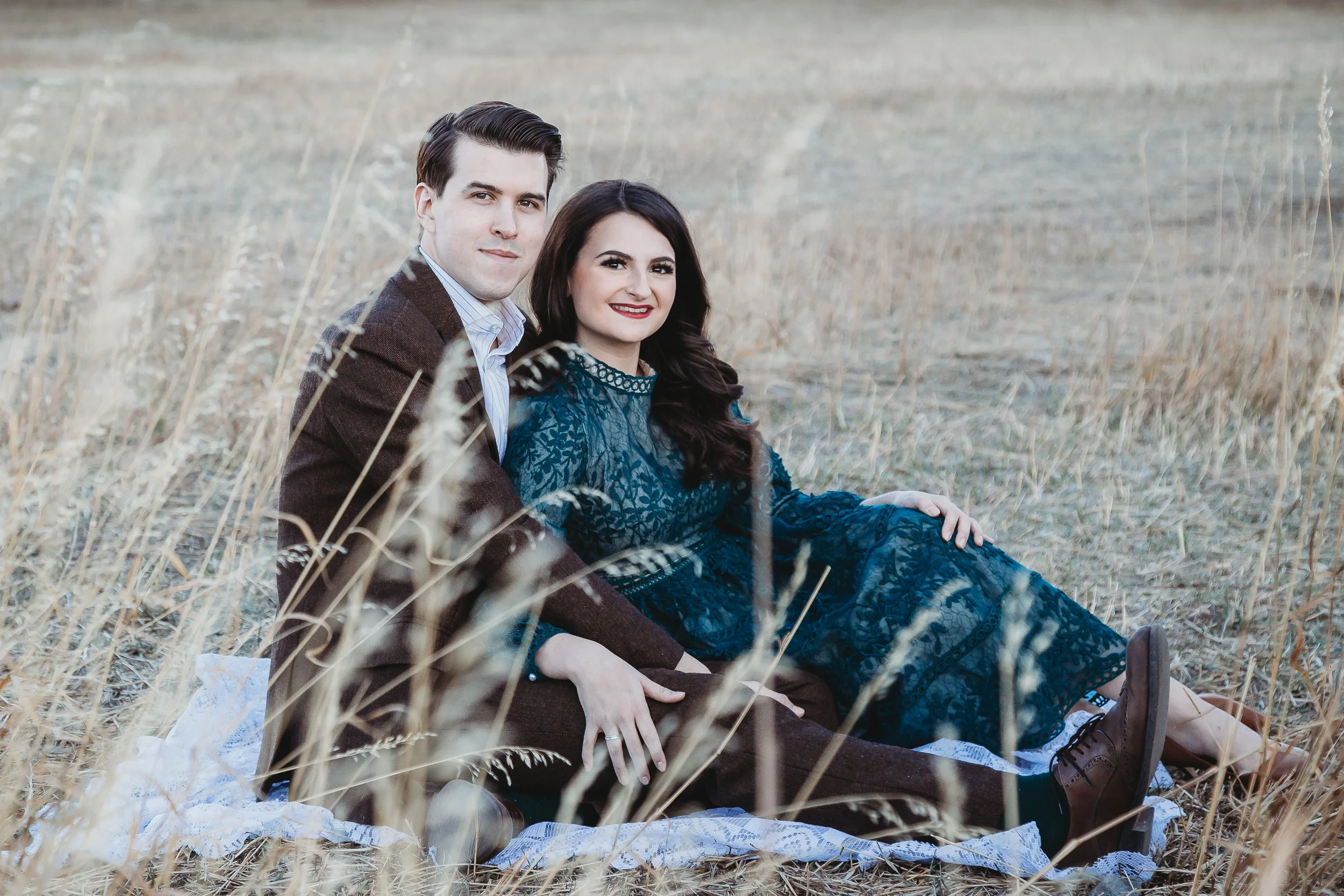 A couple sitting on a blanket in a field of tall, dry grass during daytime. The man is wearing a brown jacket and the woman is in a blue dress. They are smiling and looking at the camera.