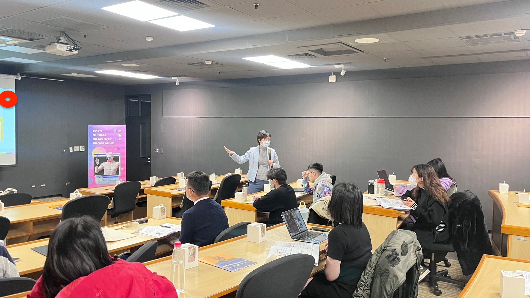 A woman presenter wearing a mask, gray blazer, and gray sweater is speaking into a microphone in a classroom or conference room. Several attendees, also wearing masks, are seated at wooden tables, some with laptops, papers, and beverages. The room has dark gray walls, ceiling lights, and a projector. A colorful poster for a 2023 global graduate program is visible in the background.