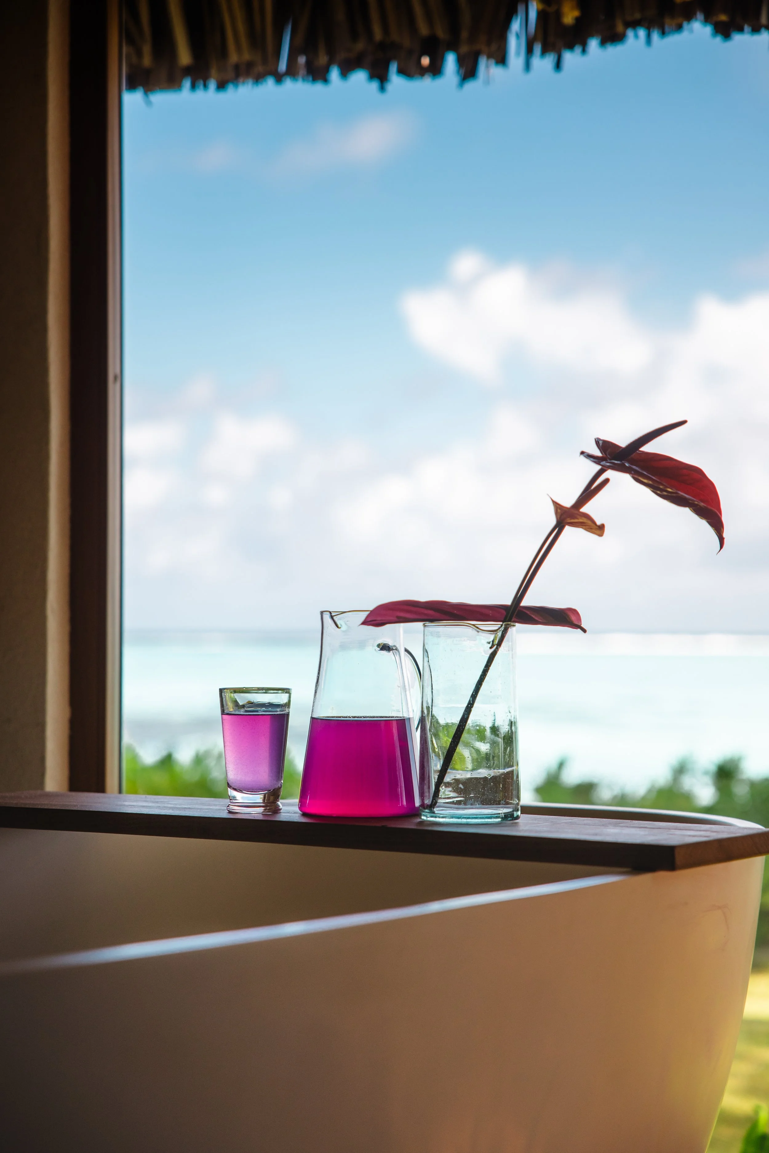 Verres et carafe avec une boisson rose sur un rebord près d'une fenêtre, avec un fond de ciel bleu et de la mer, et un pissenlit rouge dans un vase transparent.