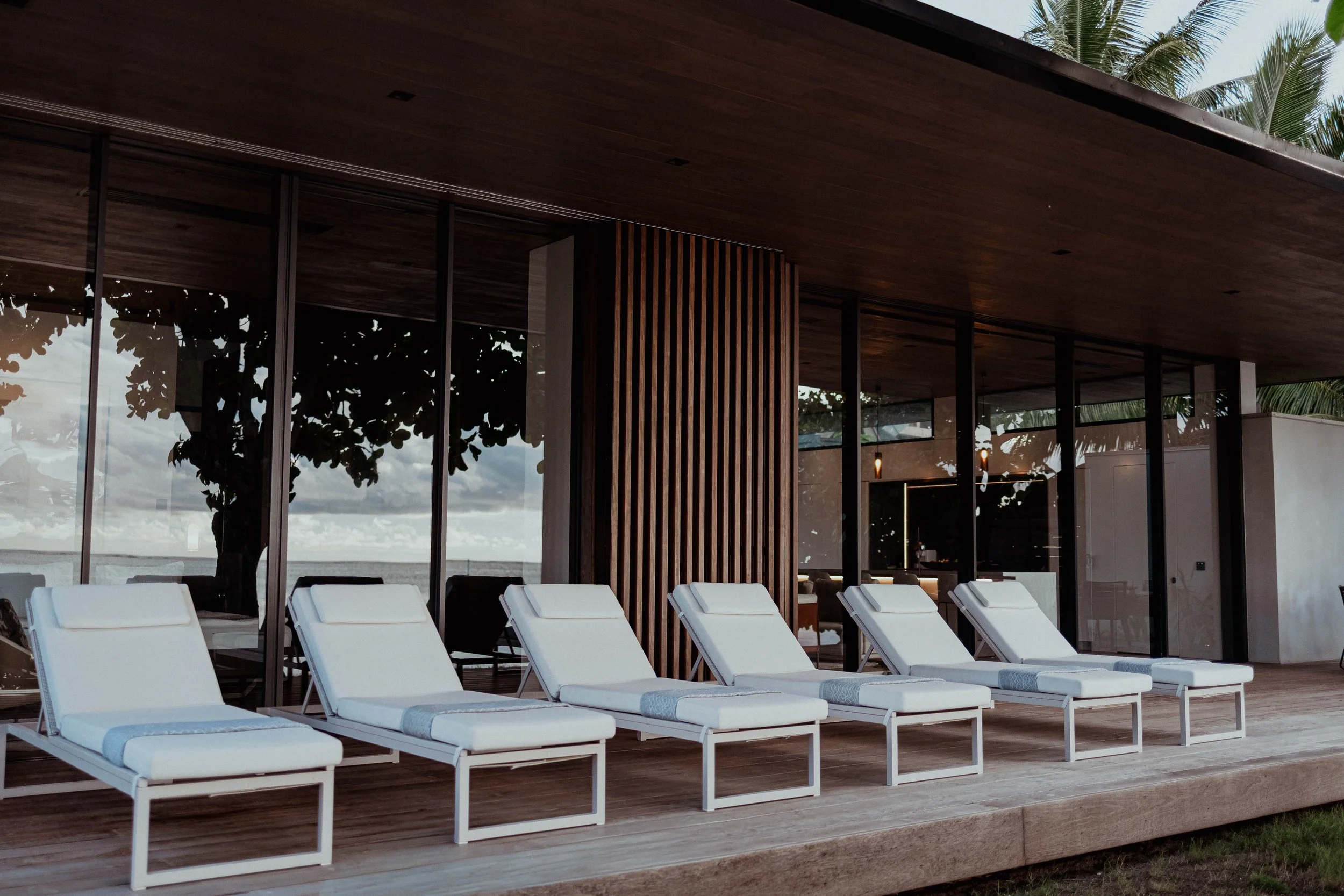 Chaises longues blanches sur une terrasse en bois devant une maison en verre avec vue sur la mer et des arbres