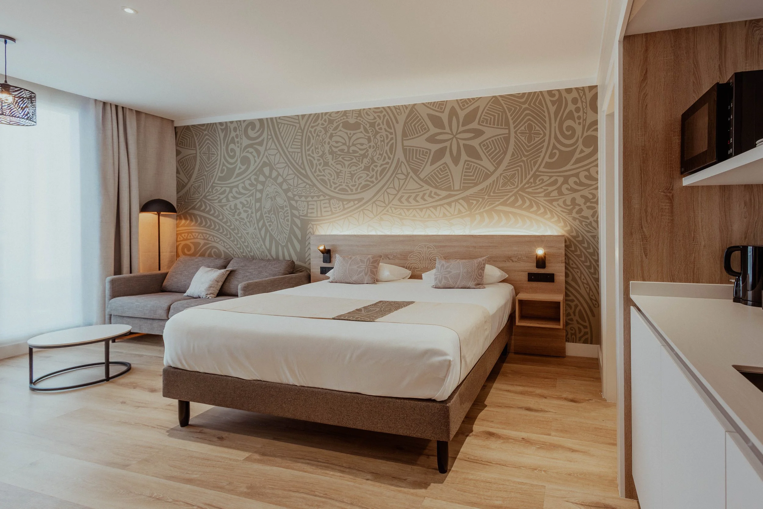Chambre d'hôtel moderne avec grand lit, mur en bois, canapé gris, table basse, mur en papier peint avec motifs polynésiens, lampes de lecture, coin cuisine avec micro-ondes et bouilloire.