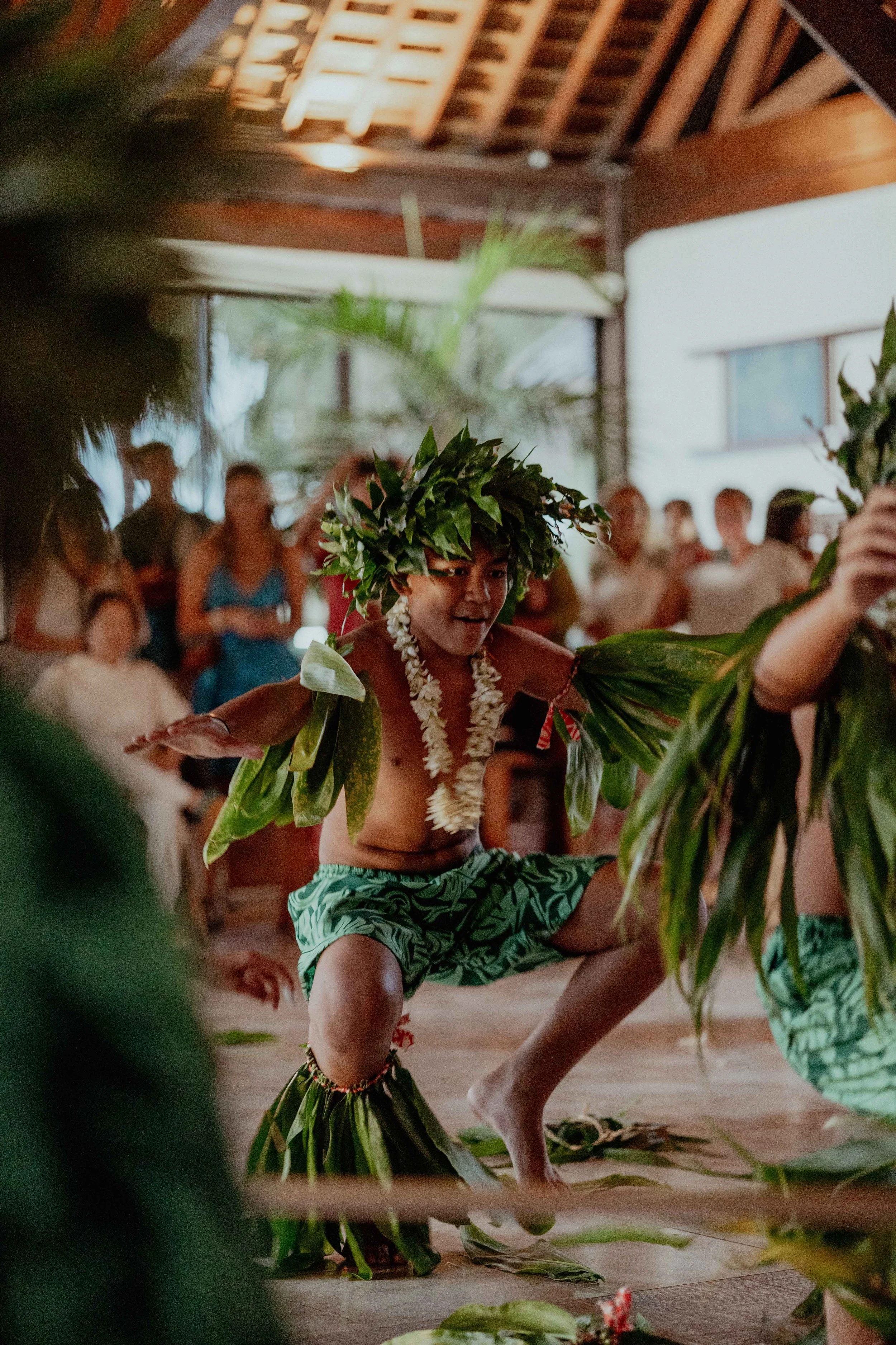 Un jeune homme danse lors d'une cérémonie traditionnelle polynésienne, portant un collier de fleurs et une couronne verte, habillé en paréo vert, avec des spectateurs en arrière-plan.