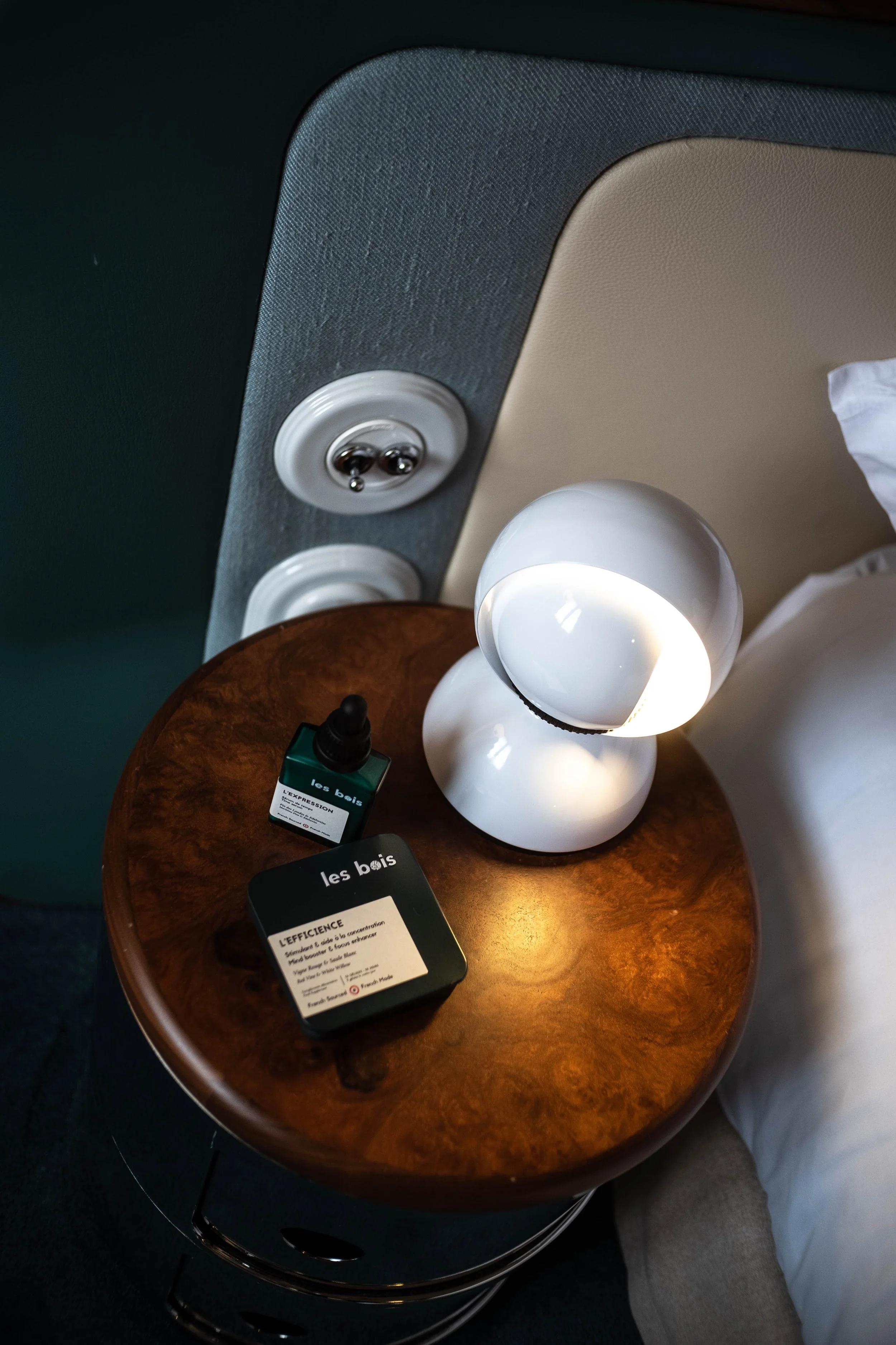Une table de nuit en bois avec une lampe blanche, une bouteille d'huile ou de parfum et une boîte en métal noire, à côté d'un lit avec une tête de lit en tissu gris et beige, près d'une prise électrique 