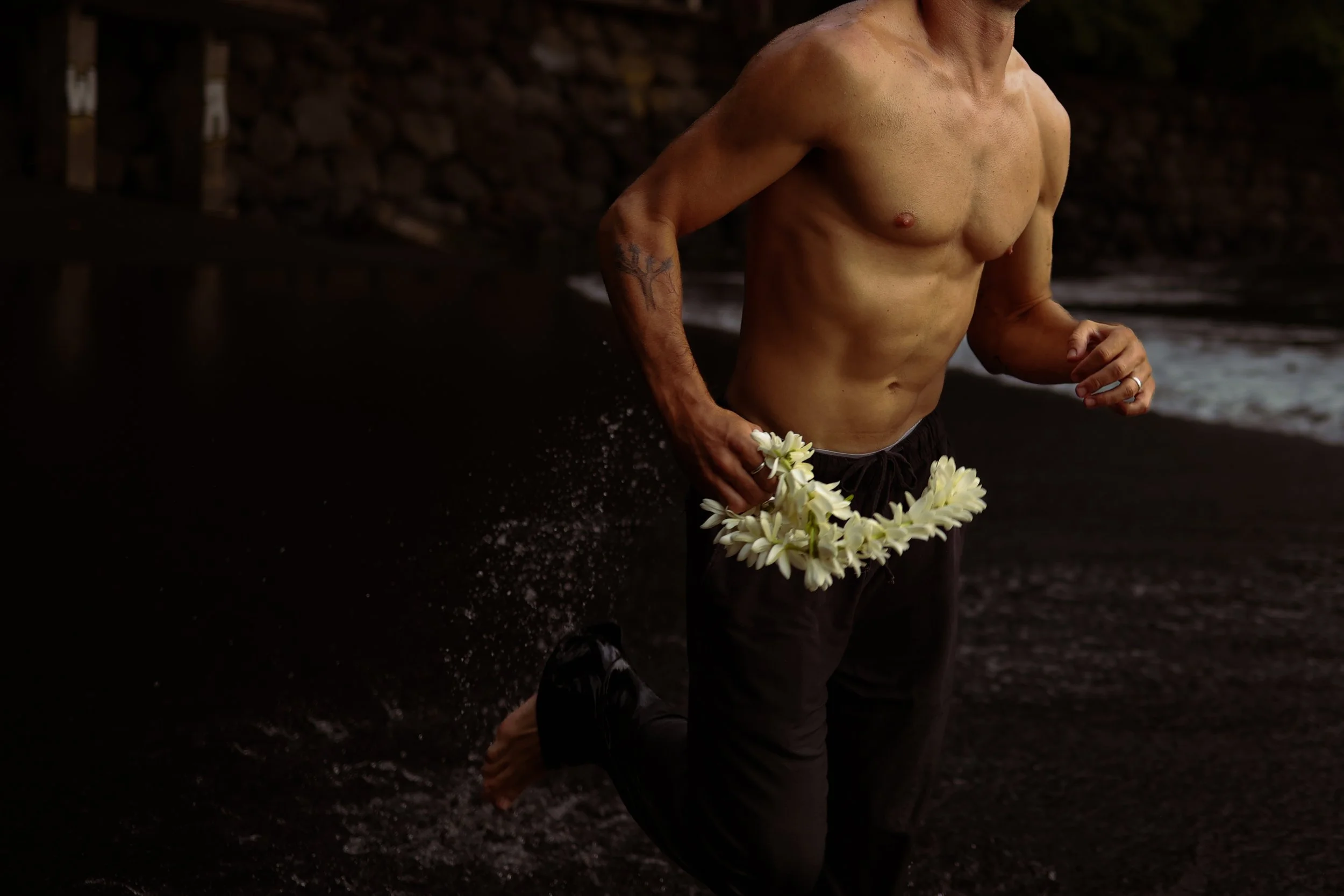 Un homme nu court sur une plage sombre, tenant un collier de fleurs blanches.