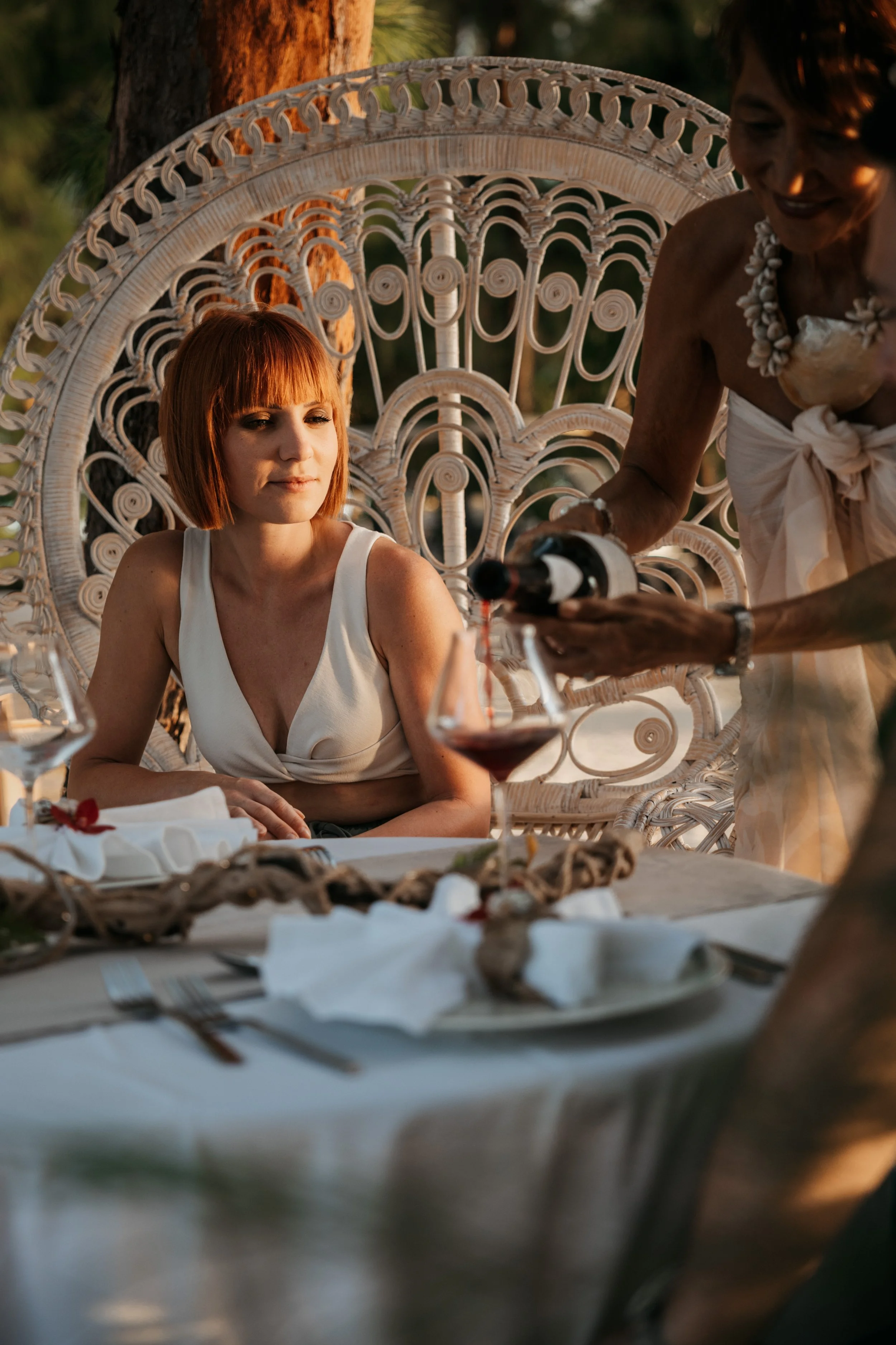 Une femme aux cheveux courts et roux assise à une table lors d'un repas, pendant qu'une autre femme verse du vin rouge dans son verre, en plein air, avec un décor naturel en arrière-plan.