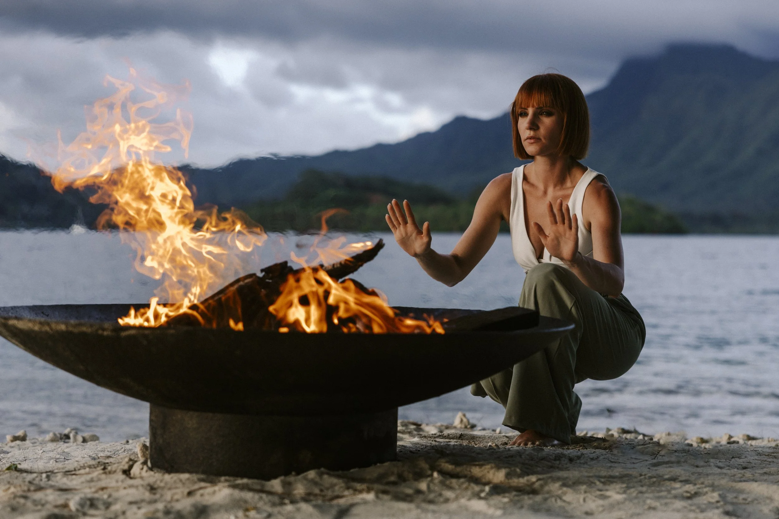 Une femme assise en face d'un feu de camp près d'une rivière, avec des montagnes en arrière-plan, lors d'une soirée nuageuse.