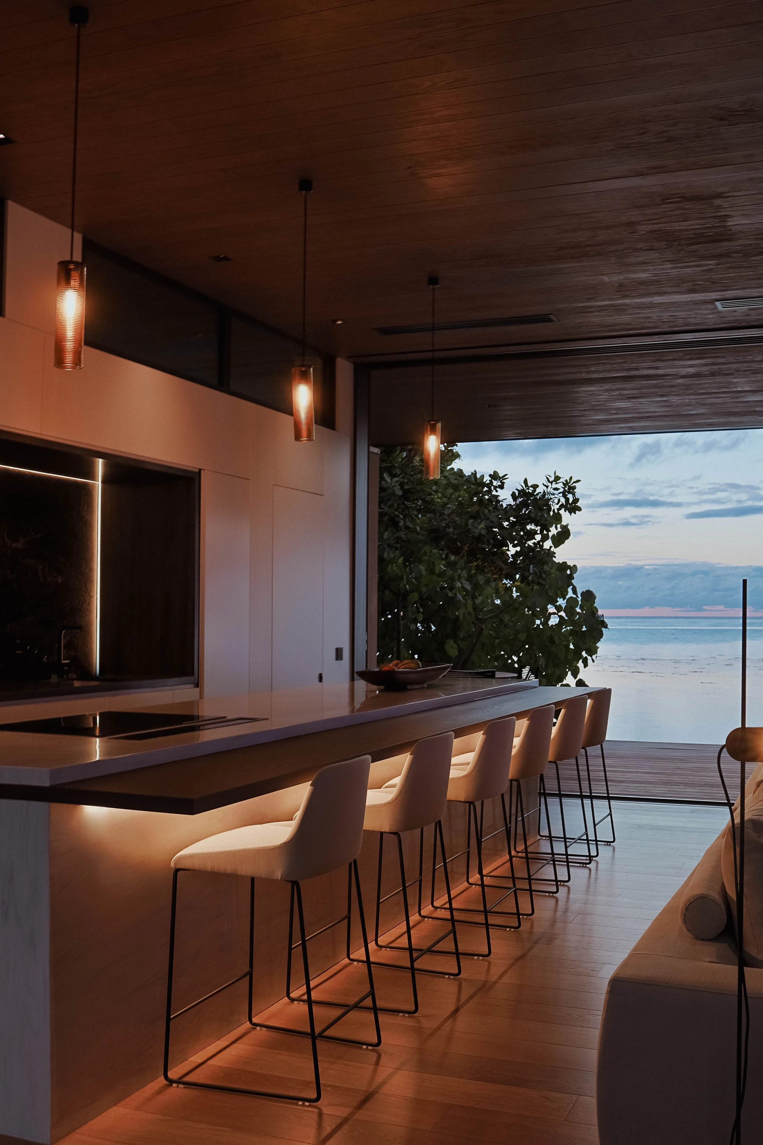 Intérieur moderne d'une cuisine avec un bar, des chaises blanches, un éclairage tamisé et une vue sur la mer au coucher du soleil.