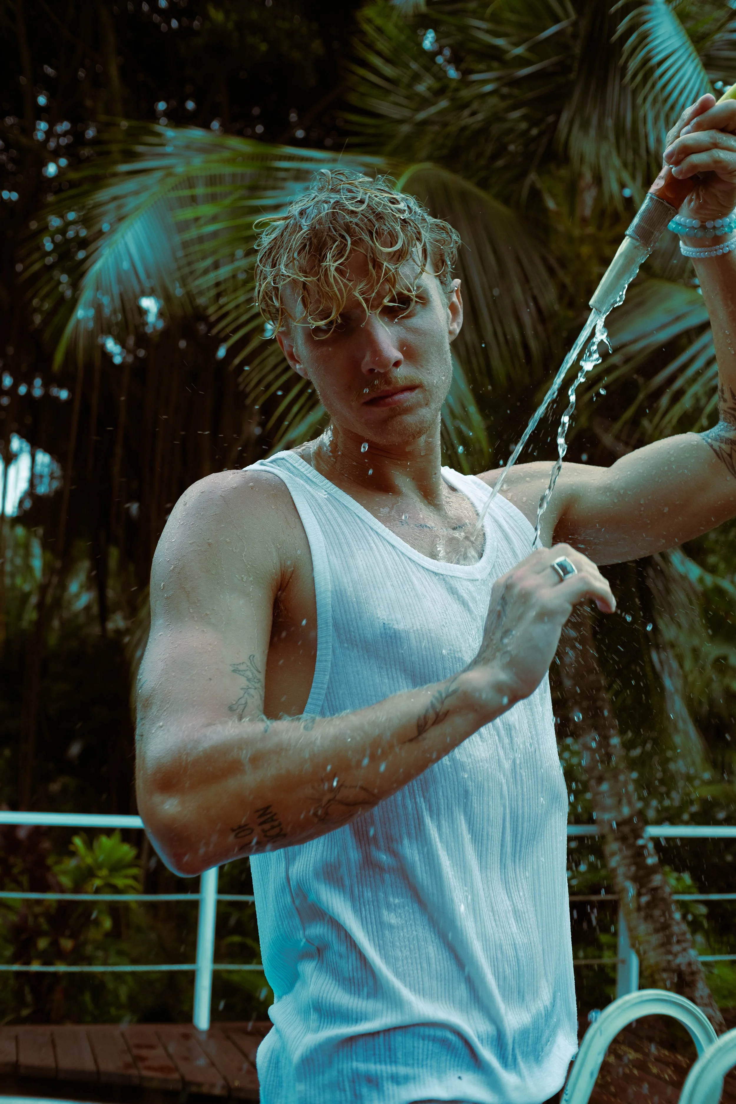 Un homme portant un débardeur blanc, tenant un tuyau d'arrosage, sous une pluie d'eau, avec des palmiers en arrière-plan.