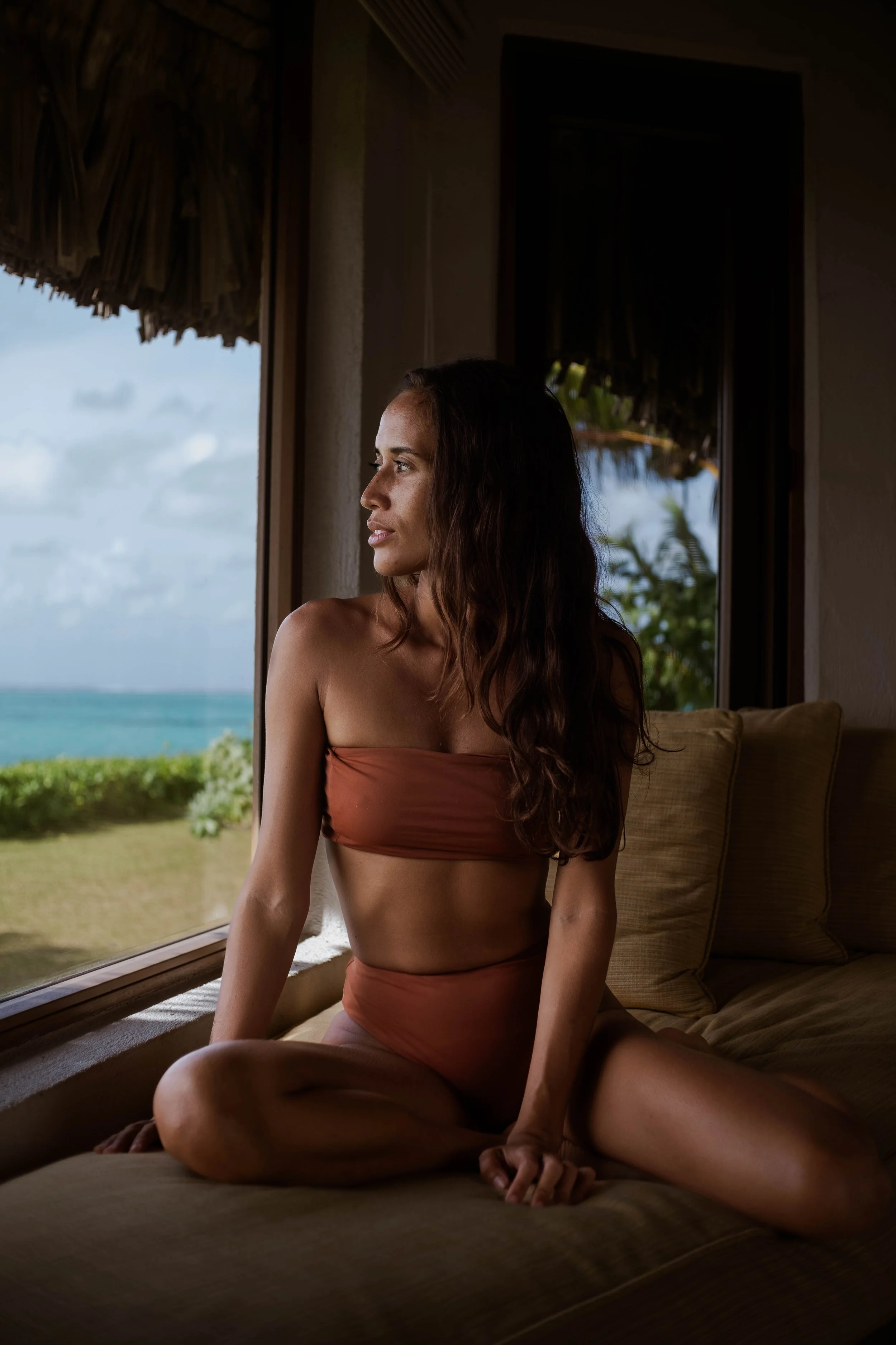 Femme assise sur un canapé à l'intérieur, regardant par une fenêtre avec vue sur la mer et la végétation tropicale.