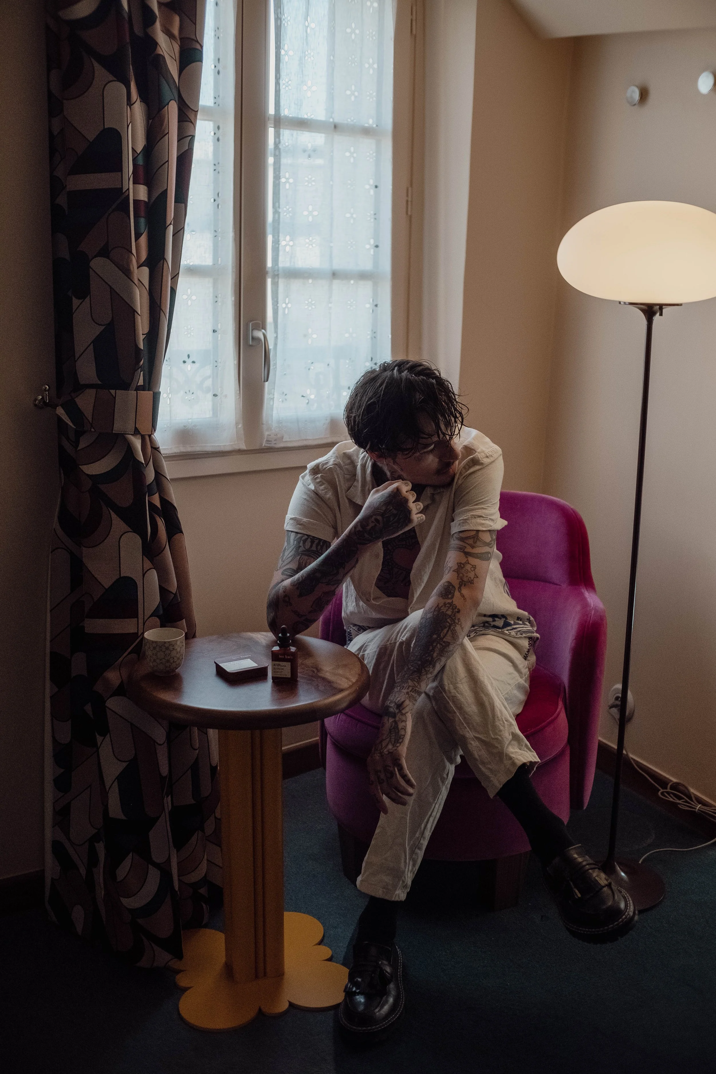 Une personne assise dans un fauteuil rose près d'une fenêtre, avec une lampe à côté, portant des vêtements clairs, avec des tatouages visibles sur les bras, tenant un objet à la main, dans une pièce avec des rideaux à motifs géométriques.