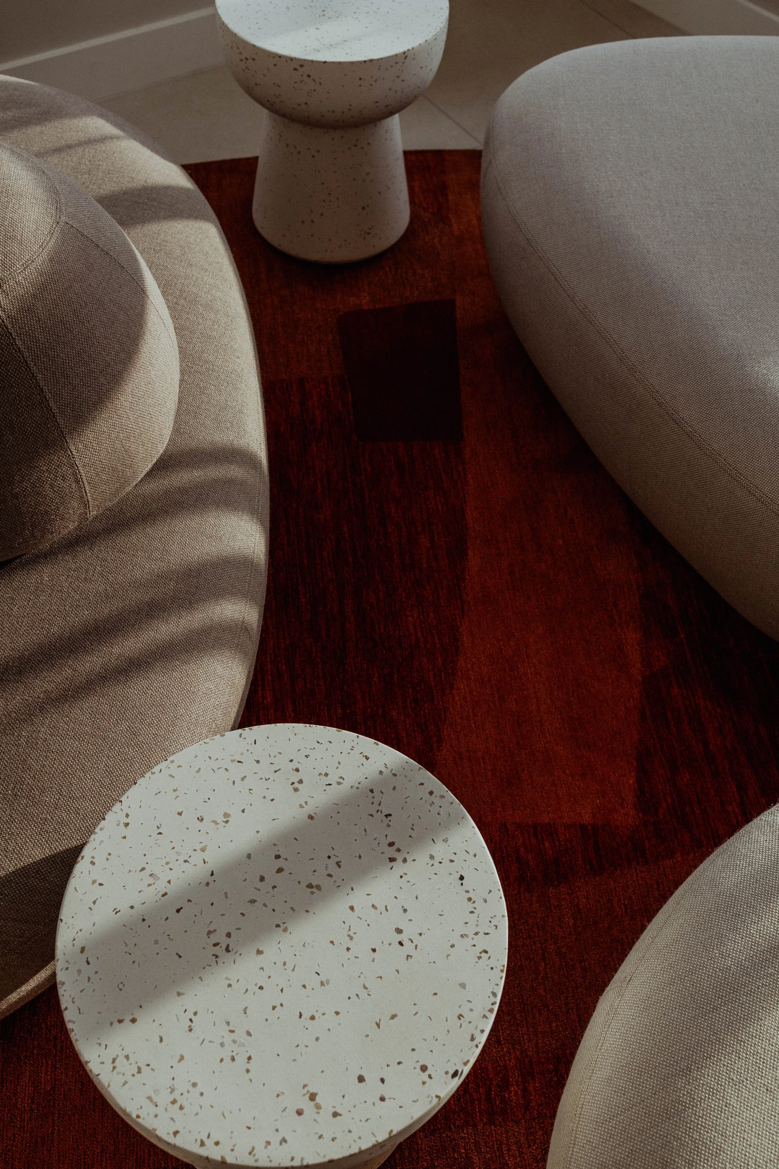 Deux tables basses en terrazzo blanc, un canapé beiges et un tapis rouge foncé dans un salon contemporain.