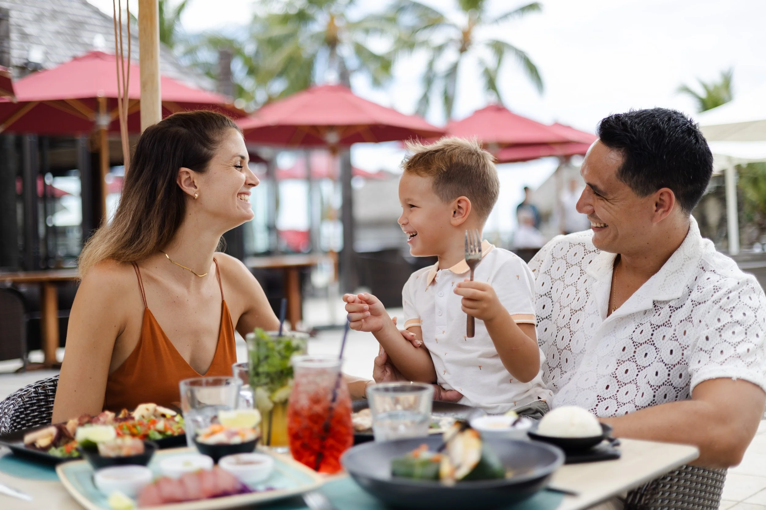 Une famille souriante partage un repas dans un restaurant en plein air avec des parasols rouges, comprenant une femme, un homme et un enfant, avec divers plats japonais sur la table.