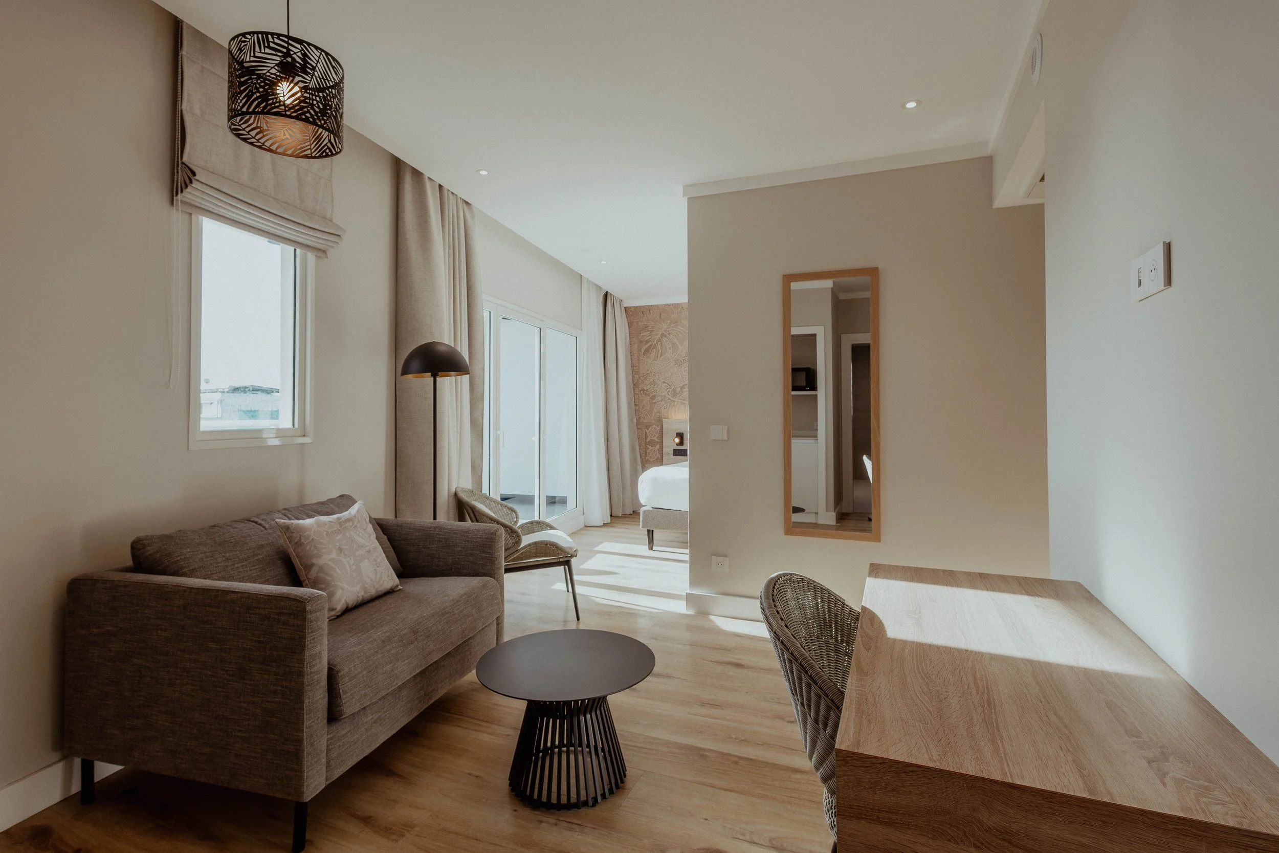 Salon moderne avec canapé gris, table basse noire, lampadaire noir, miroir mural en bois, fenêtres avec rideaux clairs, vue sur une chambre avec lit.