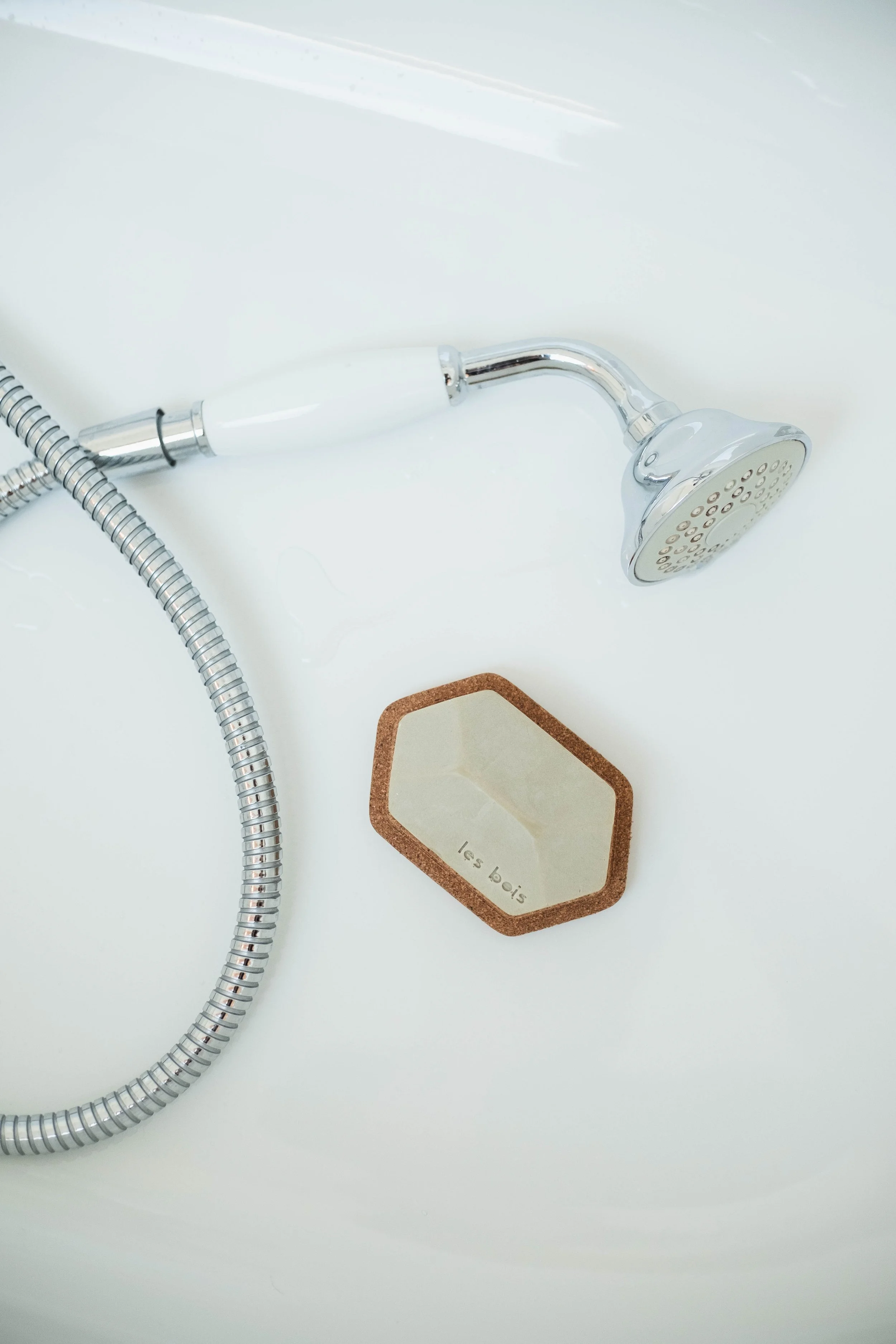 Un pommeau de douche en chrome avec un flexible métallique et un miroir de maquillage en forme hexagonale avec le texte 'les bois' écrit dessus, posé sur une surface blanche.