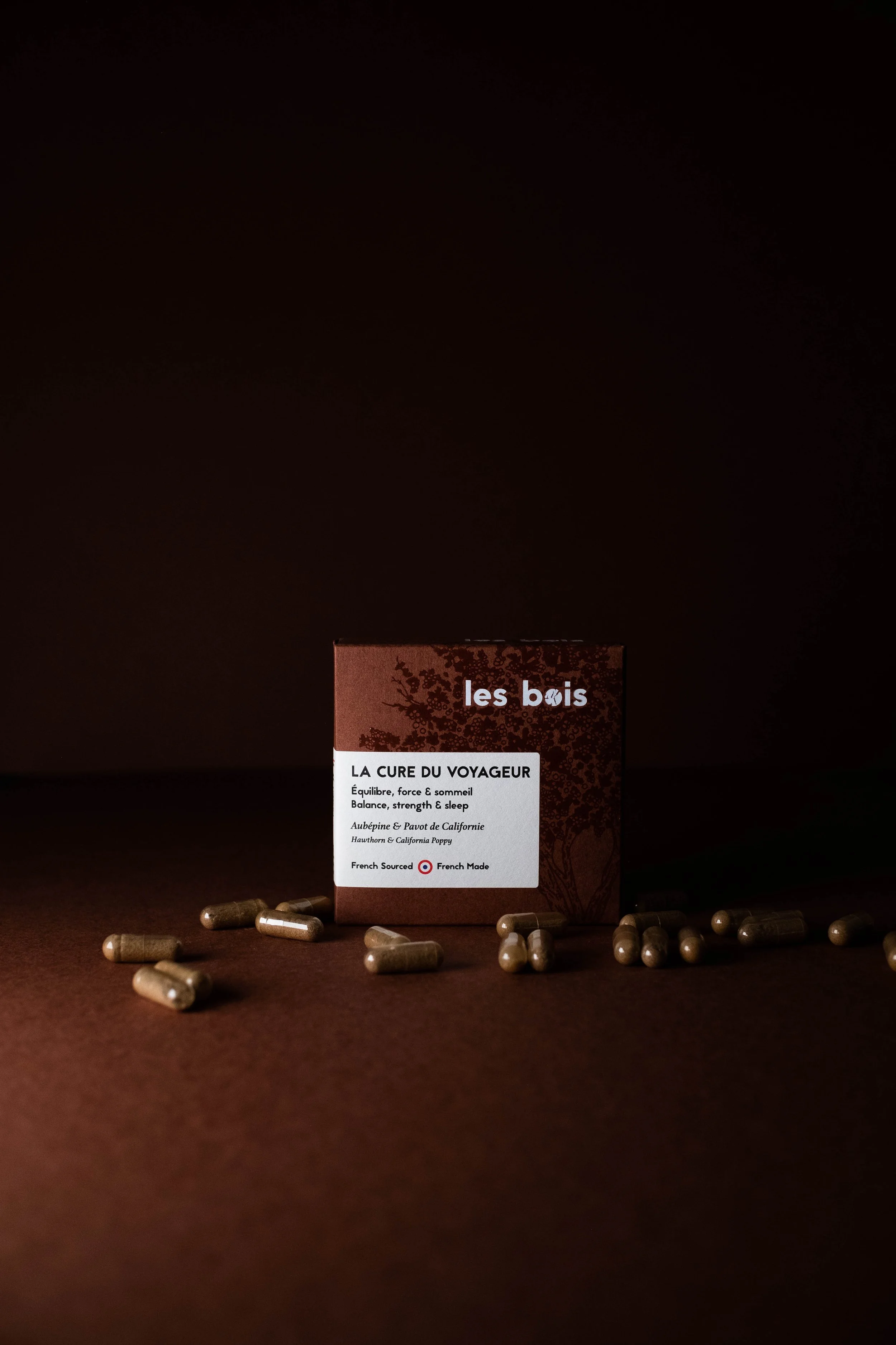Boîte de compléments alimentaires 'La Cure du Voyageur' de la marque 'Les Bois', entourée de capsules, avec une description en français et anglais sur la boîte.