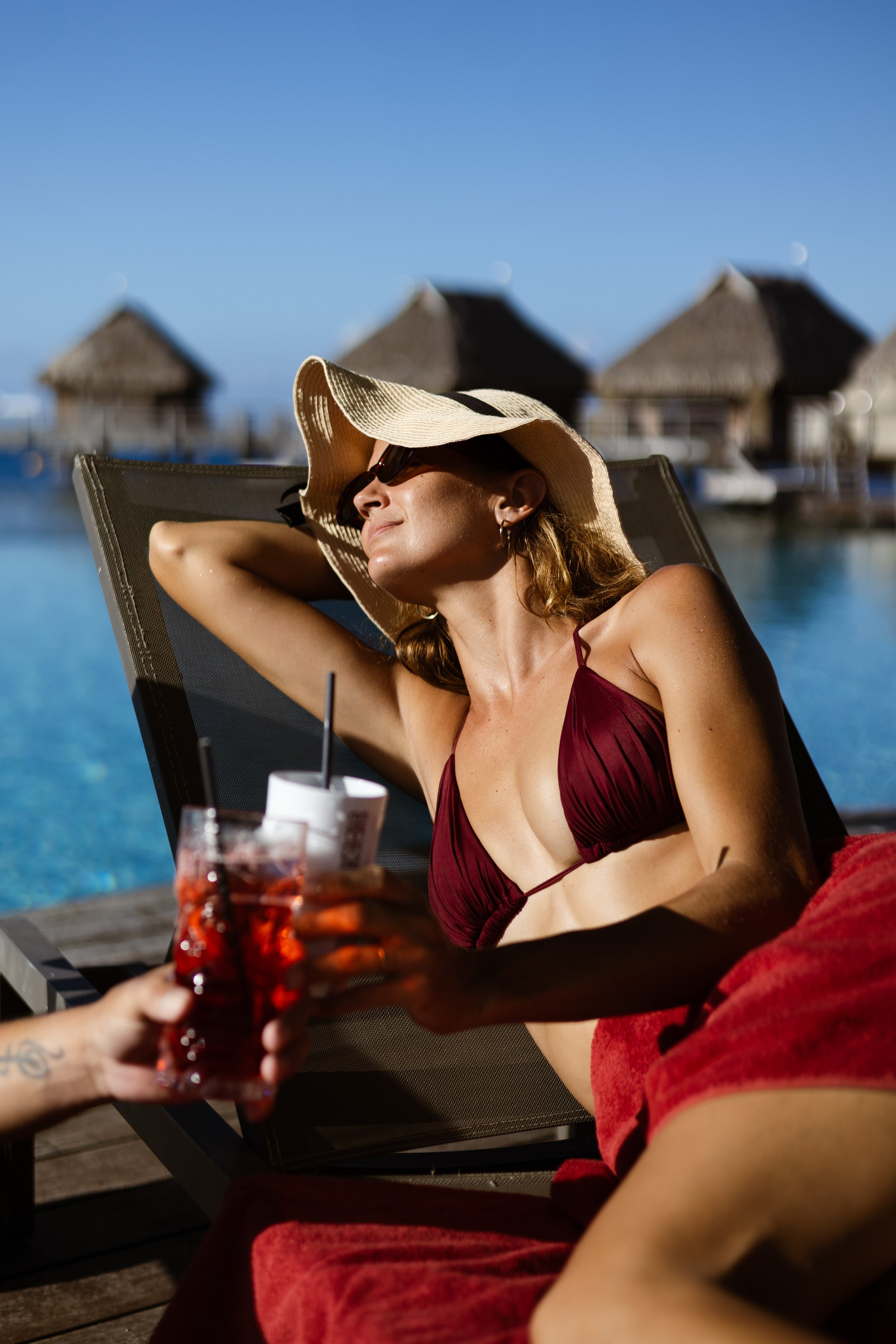 Une femme en maillot de bain rouge, portant un grand chapeau et des lunettes de soleil, profite du soleil près d'une piscine avec des cabanes en bois en arrière-plan.
