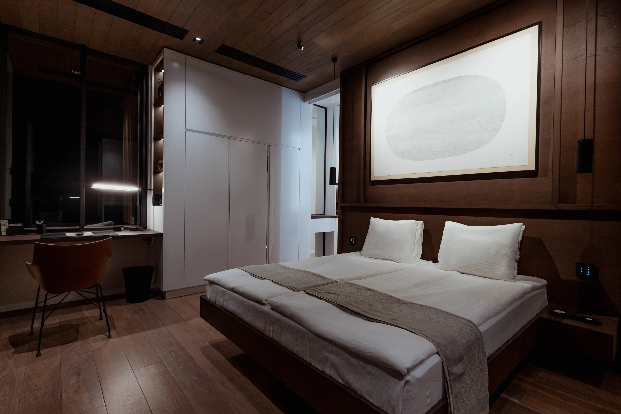 Chambre moderne avec lit double, grande peinture abstraite au mur, bureau avec chaise et lampe de bureau, placards blancs, sol en bois, ambiance chaleureuse et minimaliste.